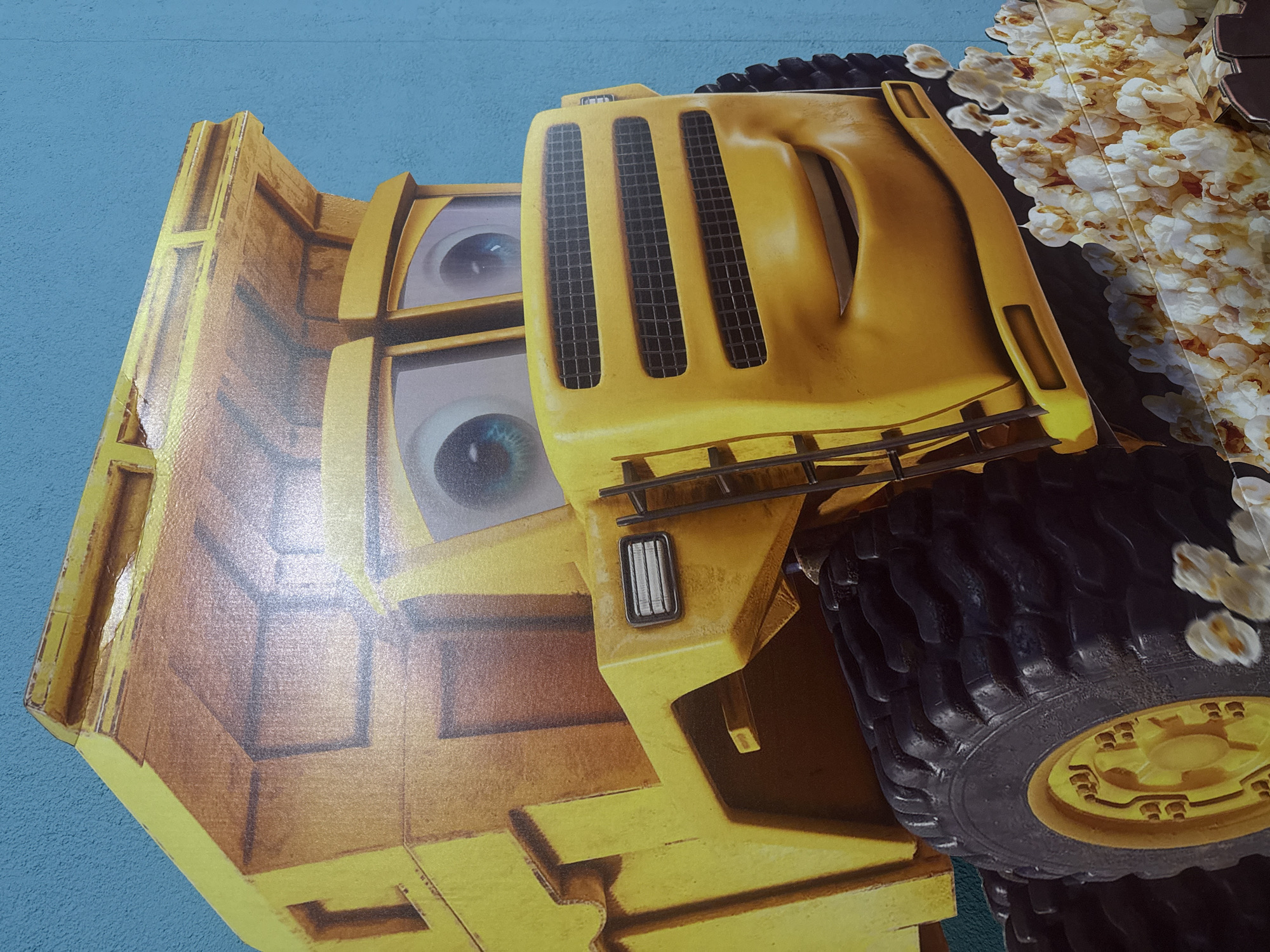 A close-up, angled shot depicts a cardboard cutout of a yellow construction vehicle, likely a dump truck. The cutout features two large eyes printed on the cab, giving it a cartoonish appearance. A large, black tire with yellow rims is partially visible on the right side of the image, and some pieces of popcorn are attached to the cutout near the tire. The background is a light blue wall with a slightly textured surface. The photo was taken in the city of Yekaterinburg, Russia.