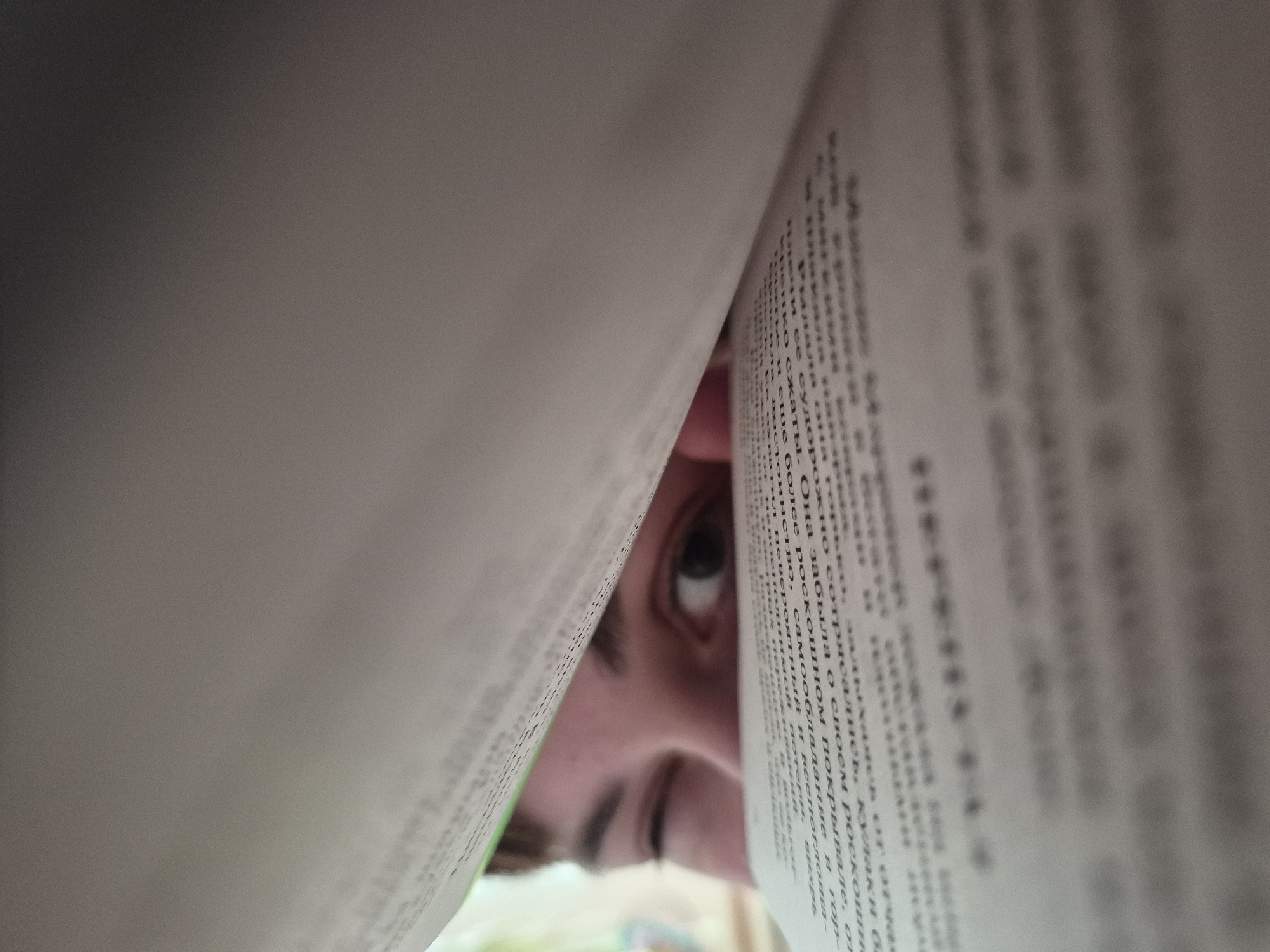 The image shows a close-up, low-angle view of a person's face peeking through the opening of a book. The book's pages are blurred, with the text on the right page partially visible. The person's eye is visible, and part of their nose and eyebrow can be discerned. The background is out of focus, revealing a hint of green.