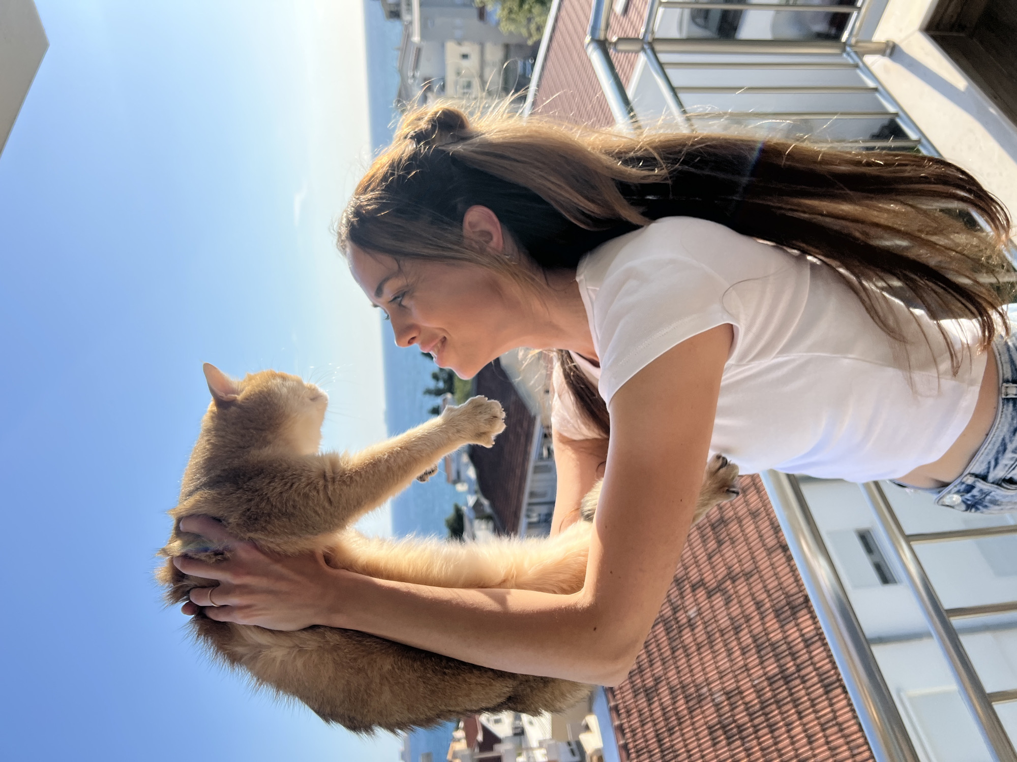 The image, rotated 90 degrees counter-clockwise from its probable intended orientation, captures a tender moment between a young woman and a cat on a sun-drenched balcony overlooking a coastal town, likely Bar, Montenegro.
If viewed upright, the young woman, with long, medium-brown hair partly styled in a half-up bun, is seen in profile, smiling and gazing affectionately down at a light brown or sandy-colored cat she holds in her arms. She wears a white short-sleeved t-shirt, and hints of blue denim overalls or jeans are visible. A thin gold ring adorns her left ring finger. The cat, with short, sleek fur, reciprocates the gaze, looking up at the woman with its front paws seemingly resting on her arm, suggesting a close bond.
The background features a vast, clear blue sky, indicating a bright and sunny day. Below the balcony, terracotta-tiled rooftops of various buildings spread across the landscape, leading to a visible expanse of the sea or ocean in the distance. A modern, silver-colored metal balcony railing frames the right side of the scene. The strong natural sunlight suggests it is daytime, casting clear shadows and enhancing the vibrant colors of the environment. The overall mood of the image is one of warmth, affection, and happiness. No visible text is present.