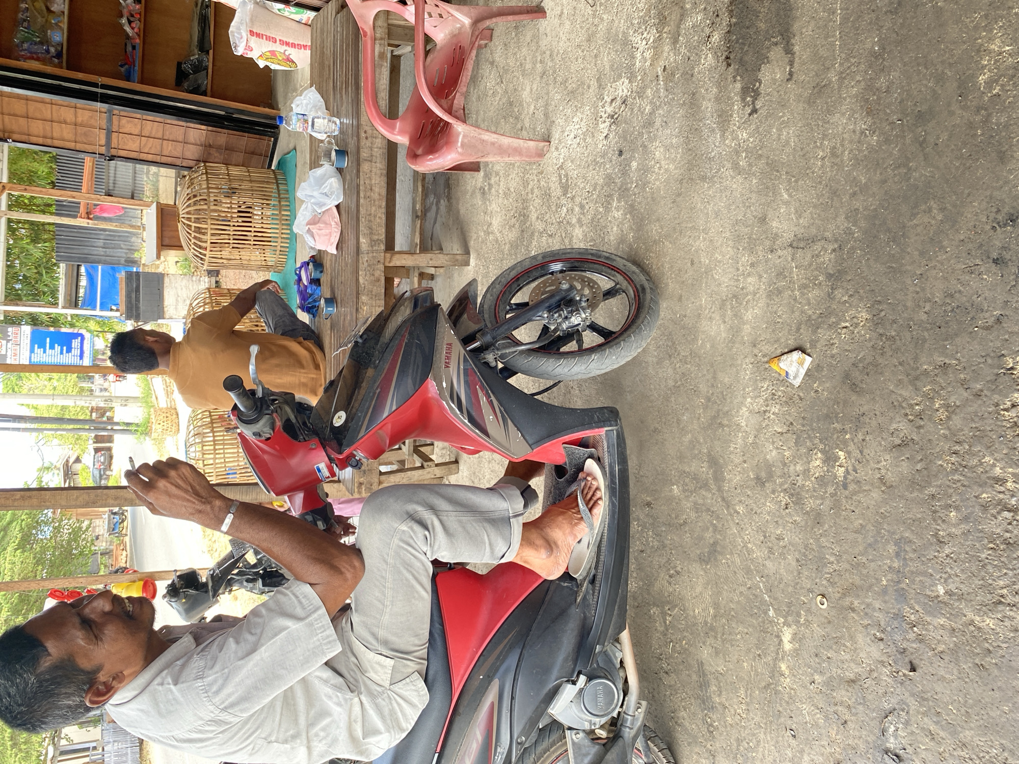 A man is sitting on a red and black Yamaha motorcycle, smoking a cigarette and wearing a light-colored short-sleeved shirt and gray pants. His right foot is resting on a flip-flop on the motorcycle's footrest. To the right, another man is sitting at a wooden table, looking away from the camera, wearing an orange shirt and gray pants. The table has several items on it, including two small cups, a pink cap, a plastic bag, and a bottle of water. Behind the table, there are two woven bird cages. The scene appears to be outdoors, possibly in a marketplace or roadside stall, with a concrete floor. In the background, there are structures made of wood and metal, and foliage. A red plastic chair is visible to the right of the table. The image was taken in the city of Bayu, Indonesia.