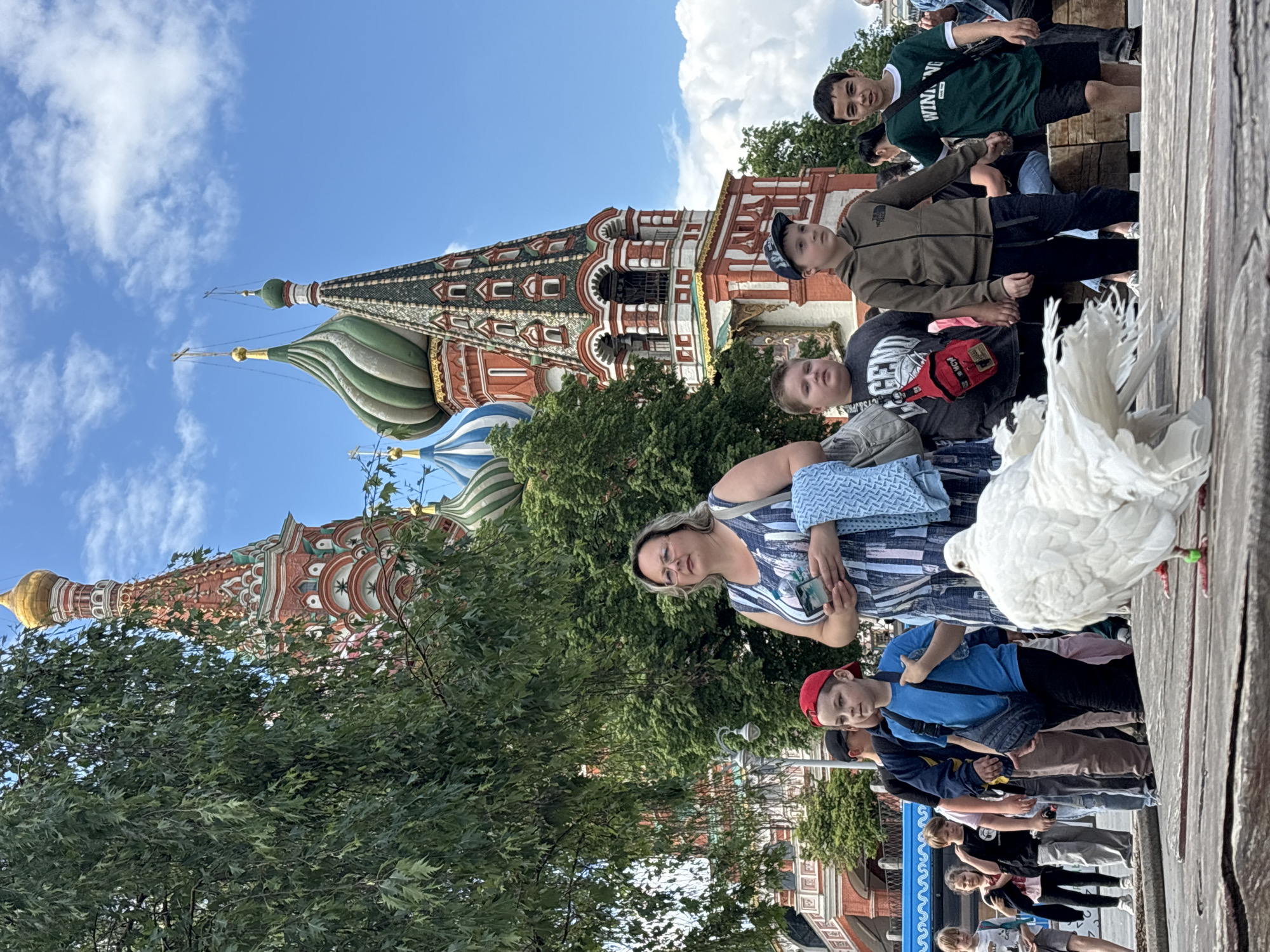 The image shows a group of people on a sunny day in Moscow, Russia, with St. Basil's Cathedral in the background. A white dove is standing on a wooden surface in the foreground, close to a woman in a blue and white striped dress who is holding a phone. Several children are positioned nearby, some looking at the camera. The cathedral, with its distinct colorful domes and spires, is partially obscured by trees. The sky is blue with scattered white clouds.