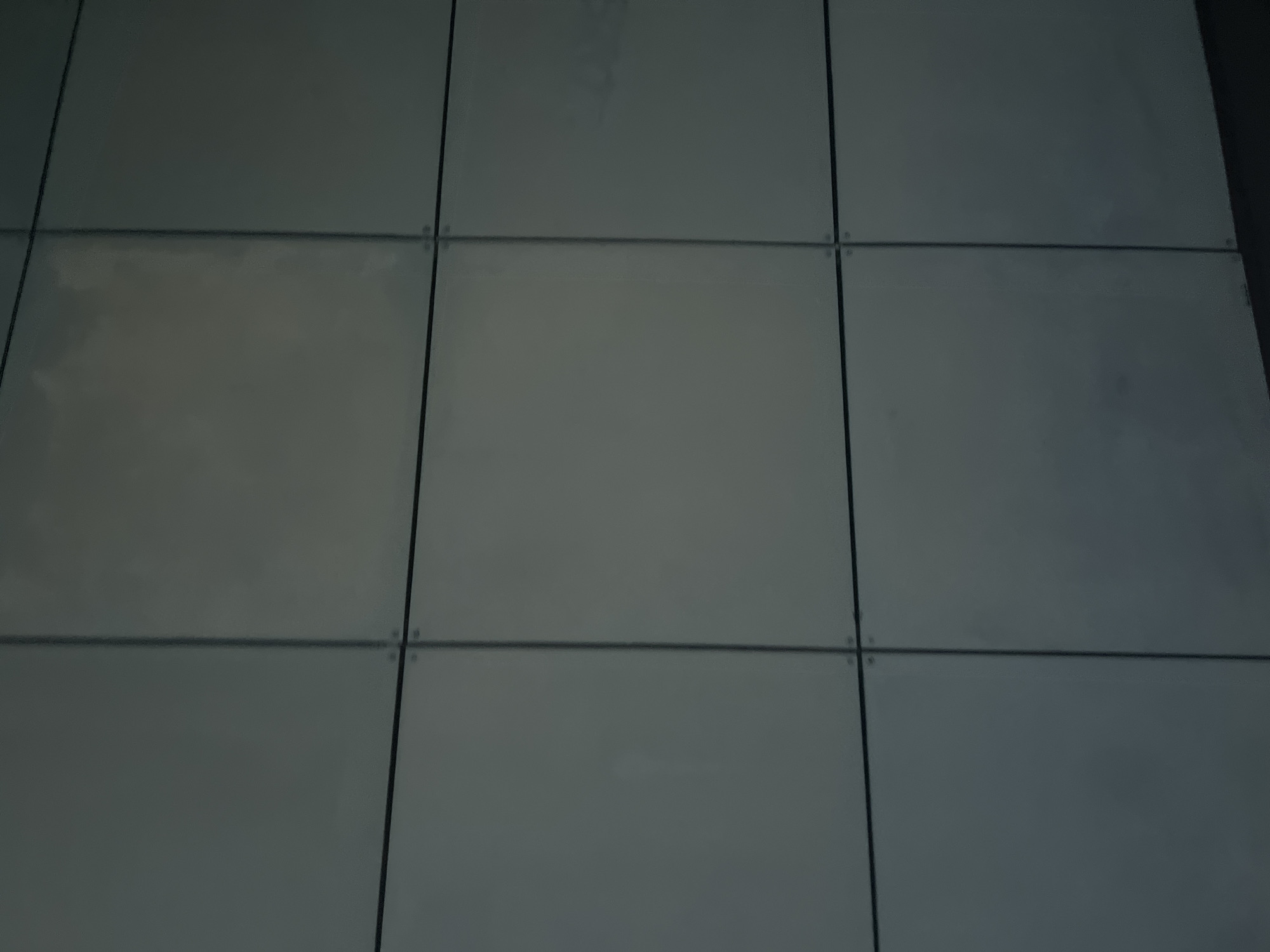 The image shows a close-up view of a building's facade, composed of a grid of nine square panels. The panels are a uniform light gray color with a matte finish and are separated by thin black lines. Each intersection of the lines appears to be secured by four small, dark-colored rivets. The building is located in Surgut, Russia. The lighting suggests it is daytime, with the light source casting a subtle, diffused glow on the surface. There are no people, objects, or discernible actions within the frame, rendering it a purely architectural detail.