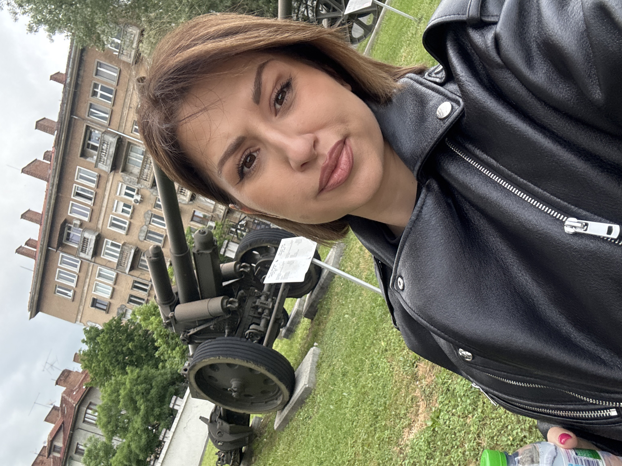 A woman poses for a selfie outdoors. The background includes a building with multiple windows and what appears to be a military anti-aircraft gun on display. The location is likely in Sofia, Bulgaria, based on the architectural style of the building and the presence of the anti-aircraft gun. The woman is wearing a black leather jacket. The setting appears to be a grassy park or museum ground. She is holding a water bottle in her right hand.