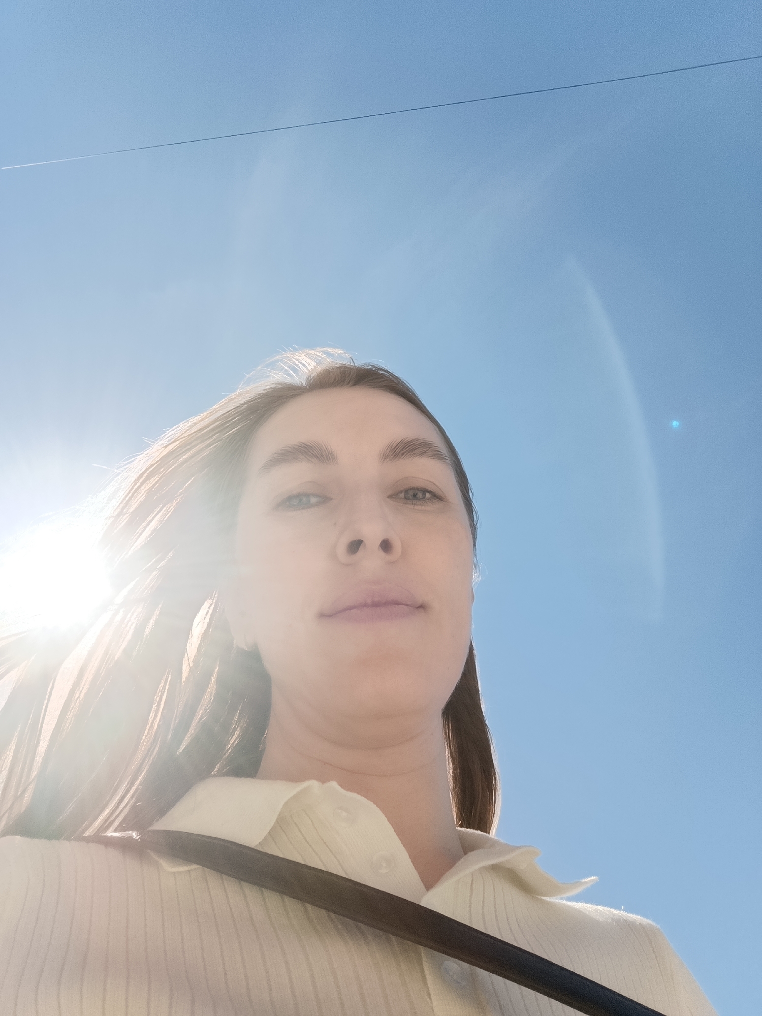 A low-angle photograph captures a woman in daylight against a clear, blue sky. She is wearing a collared, buttoned shirt, and a black strap is visible across her chest. The sun is shining brightly, causing lens flare to obscure part of her face and hair. A faint white line, potentially a contrail, stretches across the upper part of the sky. The location in the city of Chaikovsky, Russia, is not determinable from the visual information provided.