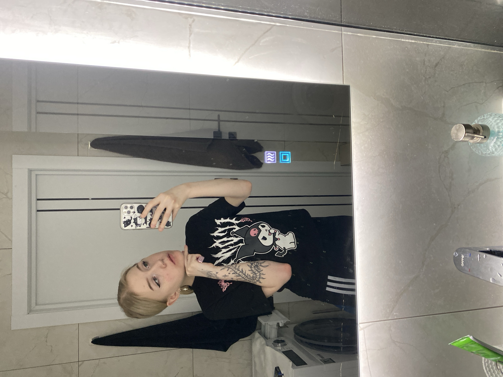 A young person with short blonde hair takes a selfie in a bathroom mirror. The person is wearing a black t-shirt with a graphic print and has tattoos on their left arm. They are holding a phone with a black and white patterned case. The bathroom features white and gray tiled walls, a white door with silver hardware, and a modern-looking mirror with illuminated touch buttons. A towel hangs on the wall behind the person. There is a washing machine partially visible in the lower part of the frame. It is not possible to determine if this photo was taken in Izhevsk, Russia.
