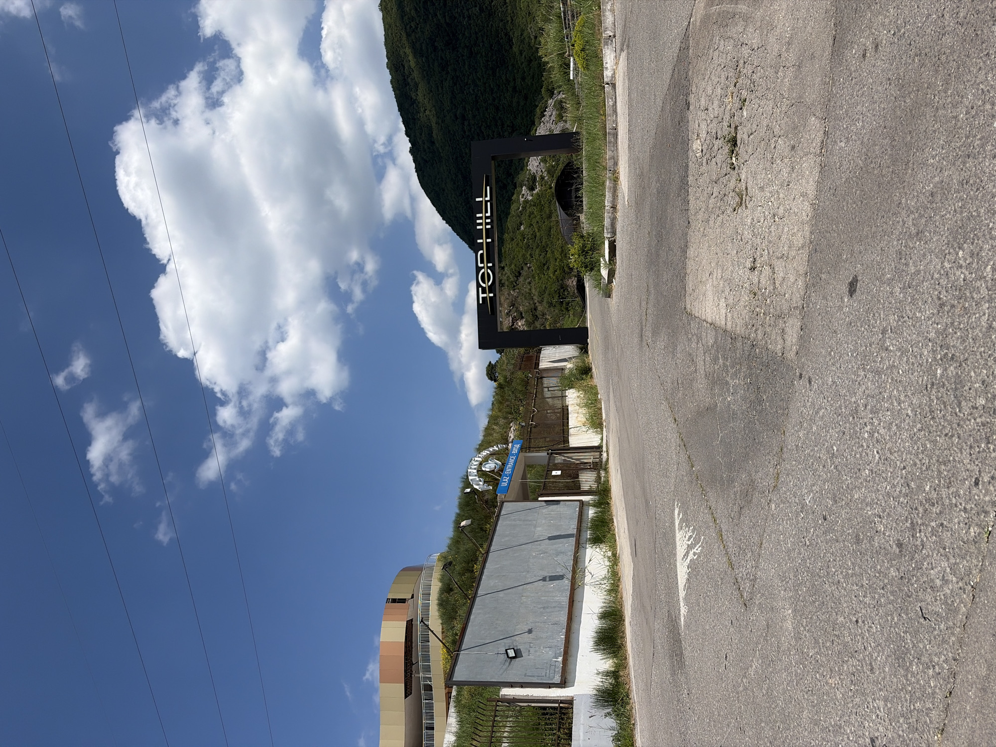 The image depicts the entrance to "Top Hill," a nightclub located in Budva, Montenegro. The entrance features a prominent black archway with "TOP HILL" written in white lettering. Beyond the archway, a curved, dark structure is visible, possibly part of the club's facilities, nestled against a lush green mountainside.
In the foreground, a paved road leads towards the entrance. To the left of the road, a large, grey, weathered billboard stands beside a fence. Further left, a multi-colored circular building, likely another part of the establishment, is situated on a grassy slope. A blue sign above a gate reads "ULAZ- ENTRANCE - ВХОД" (Entrance in Montenegrin and Russian). The sky is blue with scattered white clouds. Power lines are visible crossing the sky. The overall scene suggests a daytime view of a commercial entertainment venue in a hilly, potentially coastal, region.