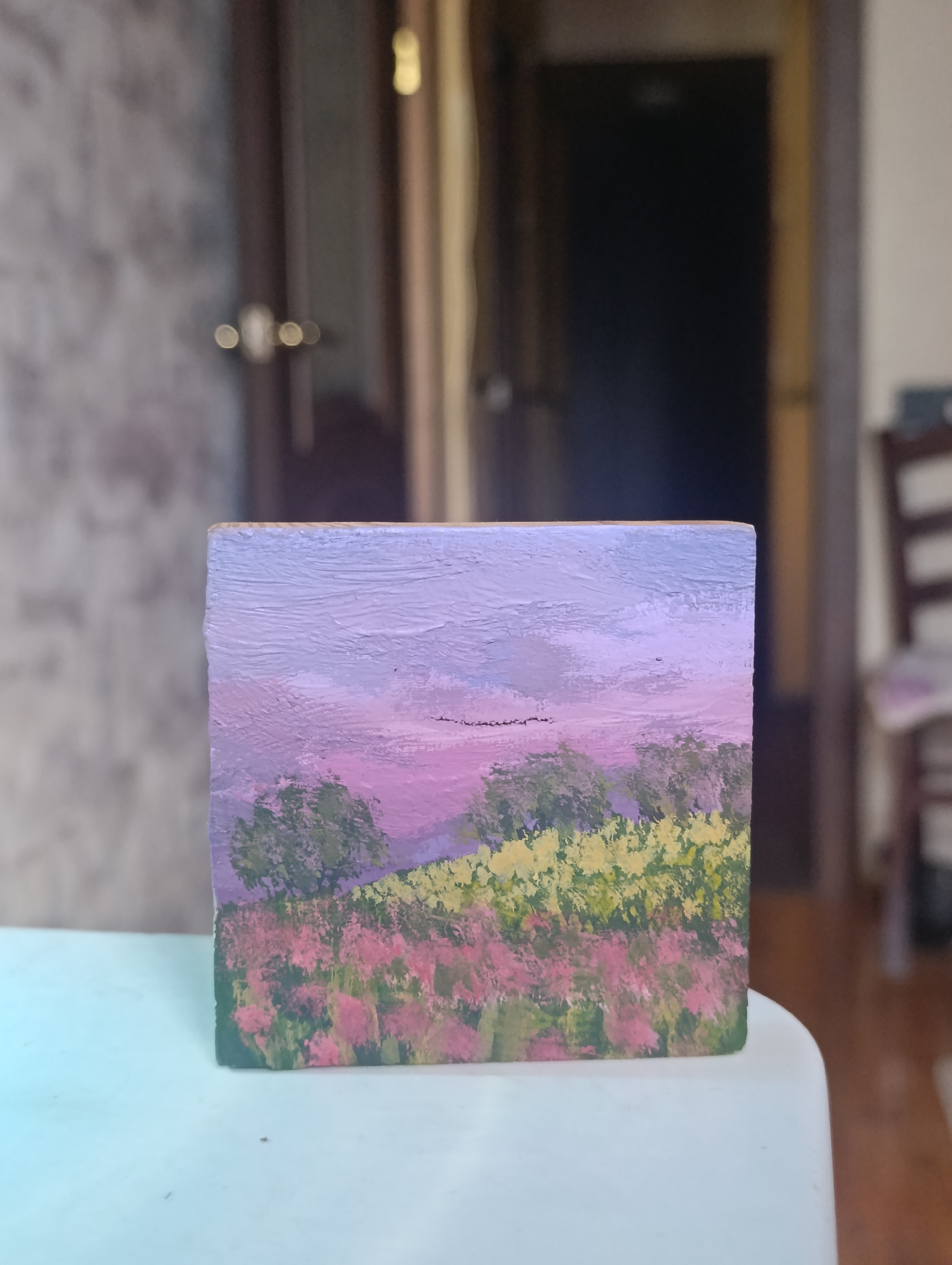 A square painted block sits on a white surface. The block depicts a landscape scene with a lavender sky, green trees, and fields of pink and yellow flowers. The scene is painted in an impressionistic style. The background is blurred and shows a doorway, a partially visible chair, and a wall. The location is impossible to determine from the image.