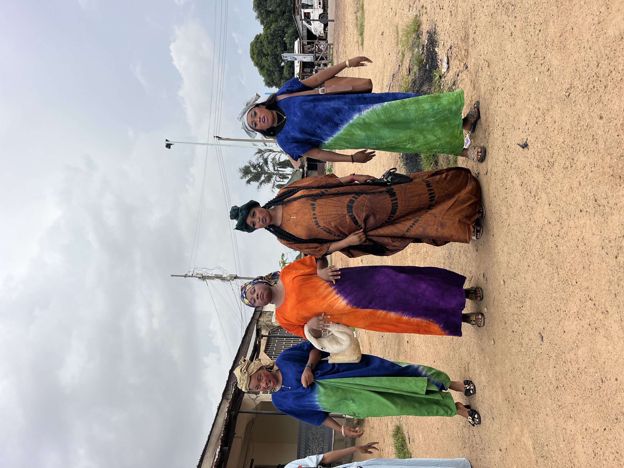 Four women stand side-by-side in an outdoor, dusty environment, possibly a village or rural area. The women are dressed in colorful, flowing tie-dye dresses. The woman on the far left wears a blue and green dress, the second woman from the left is in an orange, purple, and white tie-dye dress, the third woman from the left wears a brown and black tie-dye dress, and the woman on the far right is in a blue and green tie-dye dress. They are all wearing headscarves and makeup. The woman second from the left carries a cream-colored, textured handbag. The woman second from the right has a black crossbody bag. The woman on the far right has a brown leather shoulder bag. In the background, there are buildings with corrugated metal roofs, utility poles with wires, trees, and a white truck. The ground is covered in reddish-brown dirt. The sky is overcast with gray clouds.