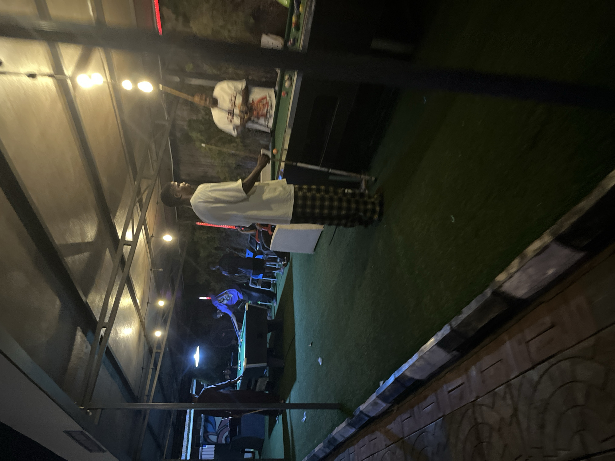 The image shows an outdoor patio area at night with artificial turf flooring and a covered ceiling. Several people are present, including two men actively engaged in playing pool at a table. One man, wearing a white t-shirt and checkered pants, is positioned to take a shot. Another man, in a white t-shirt with a graphic design, stands behind him, observing. Other individuals are visible in the background near a pool table and seating areas. The environment is lit by overhead string lights and a spotlight above the pool table. The overall scene depicts a social gathering centered around a game of pool.