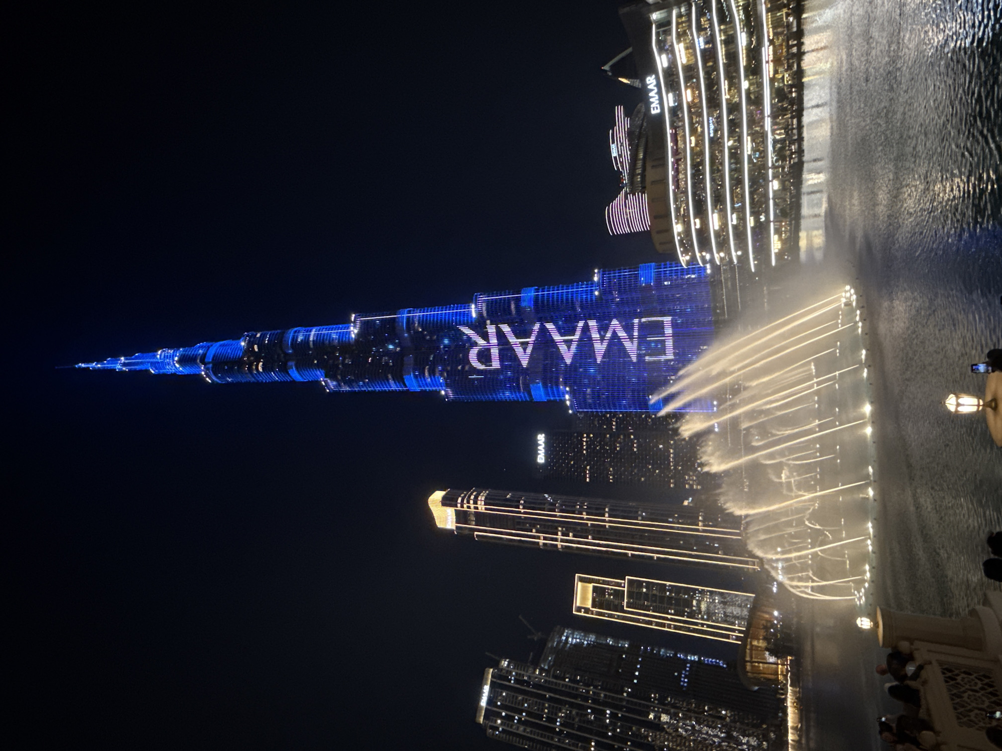 The image shows the Burj Khalifa in Dubai, United Arab Emirates, illuminated at night. The building is covered in blue lights displaying the word "EMAAR." In the foreground, the Dubai Fountain is performing with illuminated water jets. Other illuminated buildings surround the Burj Khalifa, with a dark sky in the background. People can be seen watching the fountain show.