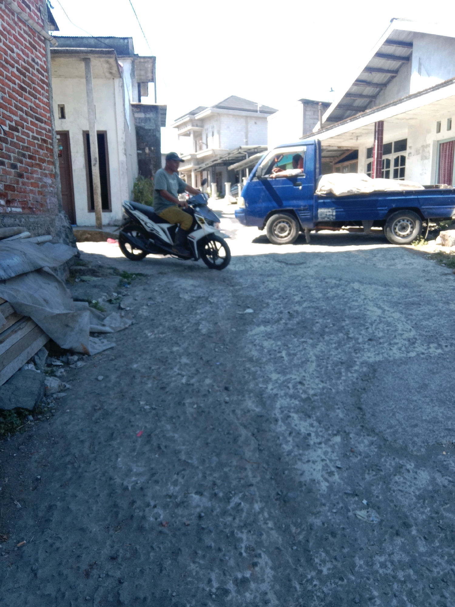 The image captures a street scene in Jurangjeru, Indonesia, under bright daylight. On the left, a person is riding a white and black scooter, wearing a light green t-shirt and khaki pants, and a dark baseball cap. In the background, a blue truck is parked on the right side of the street. The truck has its bed covered with a light-colored tarp. The street itself is made of asphalt. Buildings with white walls and corrugated metal roofs line the street. A pile of construction materials, possibly wood and tarpaulin, is visible on the left side of the road in the foreground.