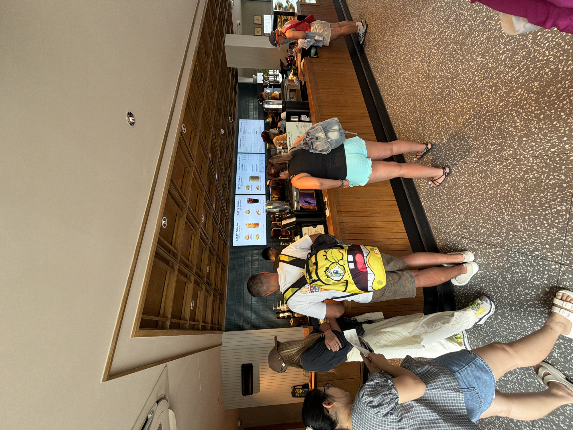 A group of people are standing in line at a Starbucks in Da Nang, Vietnam, waiting to order drinks. The interior features a modern design with a wooden ceiling and counter, and digital menu boards displaying drink options. On the left, a woman with glasses and a plaid shirt is looking at her phone. To her right, a woman wearing a baseball cap and dark shirt stands with her arms crossed, holding a brochure. A man in a white t-shirt and camouflage shorts, wearing a bright yellow Spongebob backpack, is next in line, facing the counter. Further down the counter, a woman in a black tank top and aqua shorts is looking at the menu, with a denim backpack slung over her shoulder. To her right, a woman wearing a red tank top and white shorts is standing at the counter. The floor is a speckled terrazzo pattern.