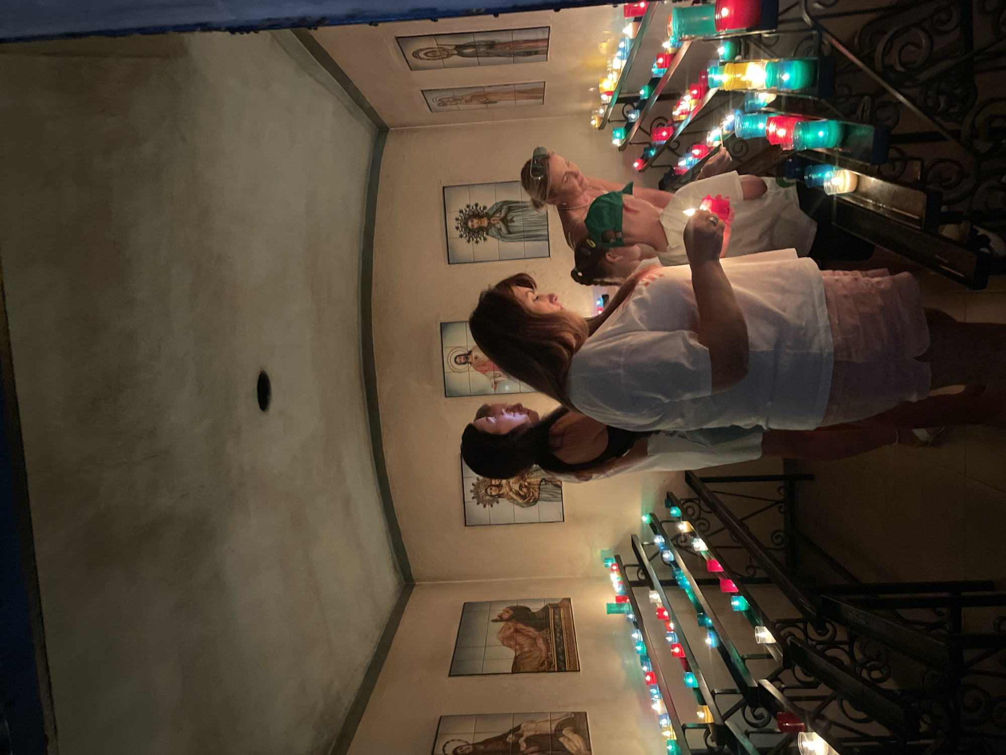 The image depicts an interior scene, likely a votive chapel, filled with numerous lit candles. Several people, including adults and a child, are present and appear to be lighting or observing the candles. The environment is dimly lit, with the primary illumination coming from the candles themselves, which are of various colors including red, green, blue, and yellow. The walls are adorned with religious imagery, specifically portraits or icons. The scene suggests a place of worship or reflection. This chapel is located in Calella, Spain, within the Sant Pere Parish Church.