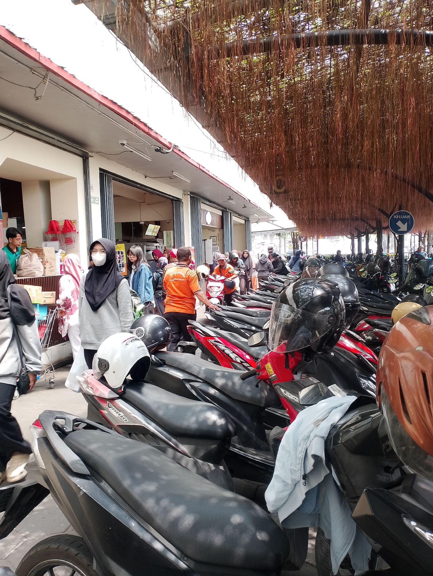 The image shows a parking lot filled with motorcycles under a canopy of hanging vines. In the foreground, several motorcycles are parked closely together, with helmets placed on some of them. A person in a grey hoodie and black hijab, wearing a white mask, stands near the motorcycles. To the left, a person in a grey jacket walks past a shopping cart and a counter area with various products. In the background, more motorcycles and people are visible. A blue sign with white text and an arrow pointing left reads "KELUAR," indicating an exit. The architecture suggests a public building or market. The presence of a sign in Indonesian and the style of motorcycles and clothing are consistent with a location in Yogyakarta, Indonesia.