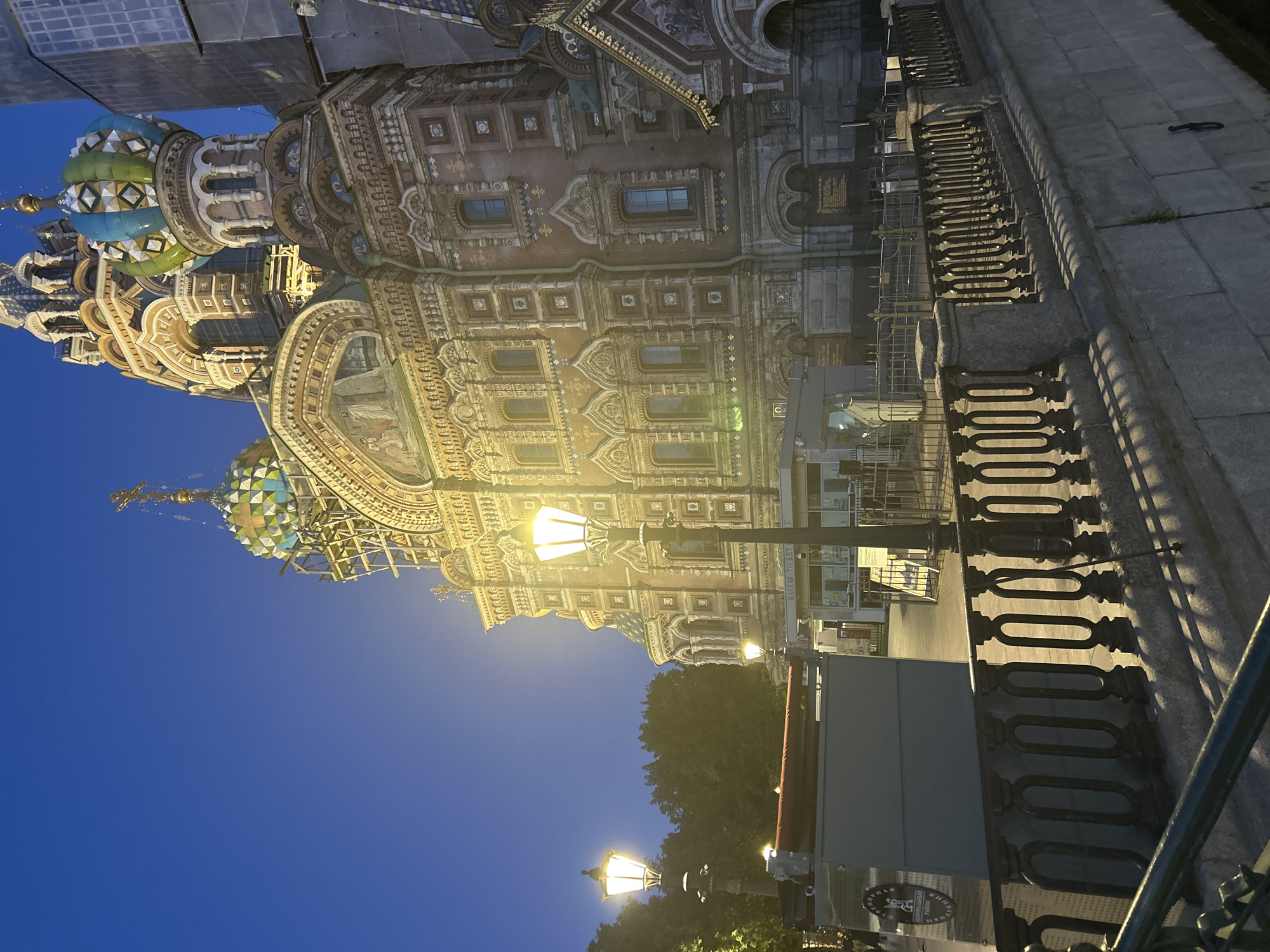 The image captures the Church of the Savior on Spilled Blood in Saint Petersburg, Russia, at dusk. The sky is a deep blue, transitioning from twilight to night. The intricately designed cathedral is illuminated by streetlights, highlighting its ornate facade with colorful domes and mosaic details. Scaffolding is visible on one of the domes, suggesting ongoing restoration or maintenance work. In the foreground, a stone embankment lined with a decorative black metal railing runs along a body of water, casting long shadows. A lamppost with a bright yellow light stands prominently on the right side of the frame. To the left, another lamppost illuminates a red awning over what appears to be a kiosk or ticket booth. Trees can be seen in the background on the left. The overall scene conveys a sense of historical grandeur and serene evening ambiance.
