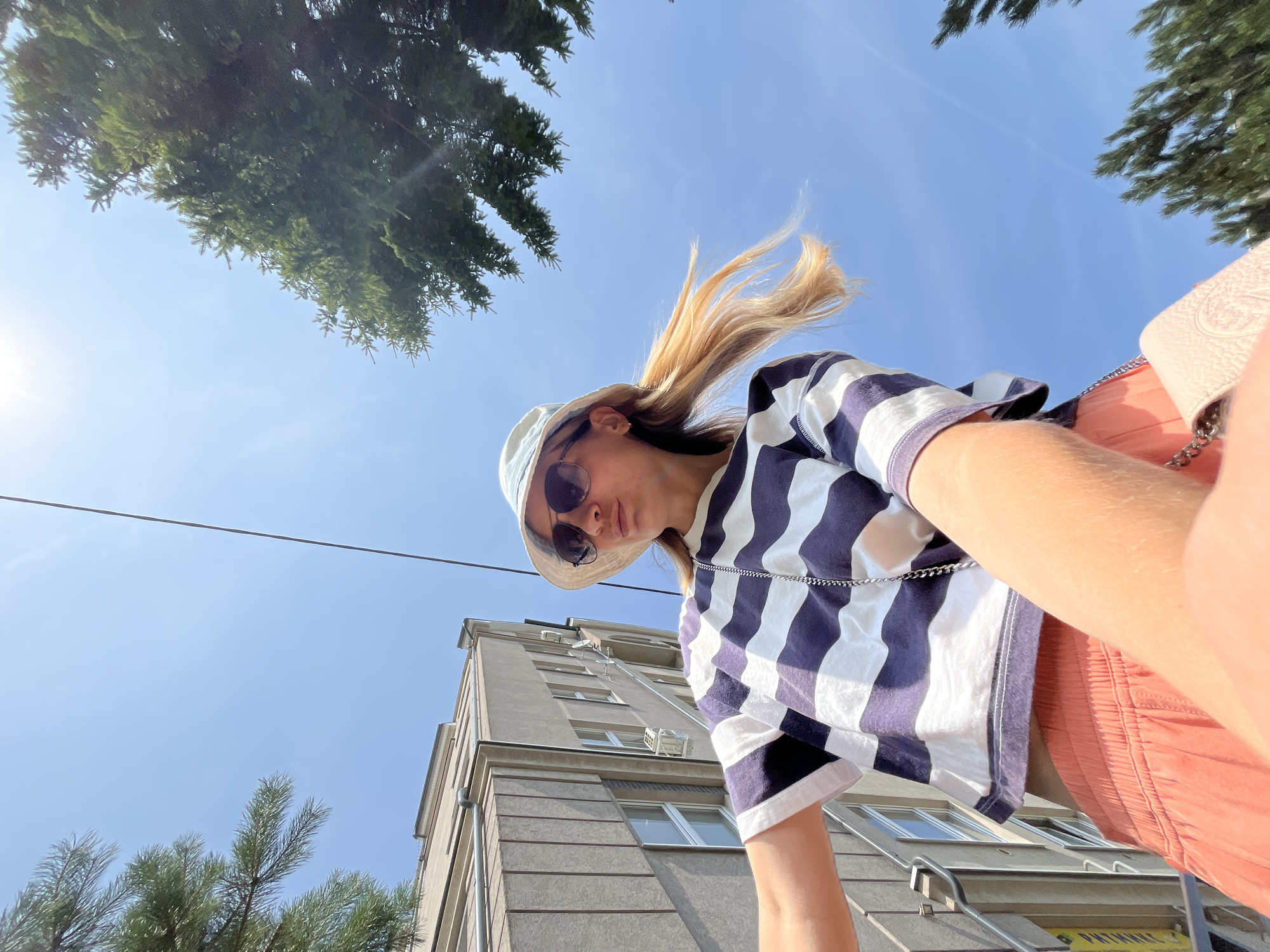 A low-angle shot captures a woman wearing a bucket hat and sunglasses, with her blonde hair blowing in the wind. She is dressed in a navy and white striped t-shirt and peach-colored shorts. She appears to be taking a selfie with her arm extended. In the background, a modern building with multiple windows stands tall, and evergreen trees frame the upper portion of the image, against a clear blue sky. The image is taken outdoors, likely in a city environment, possibly Pionersky, Russia, given the presence of urban architecture and vegetation.