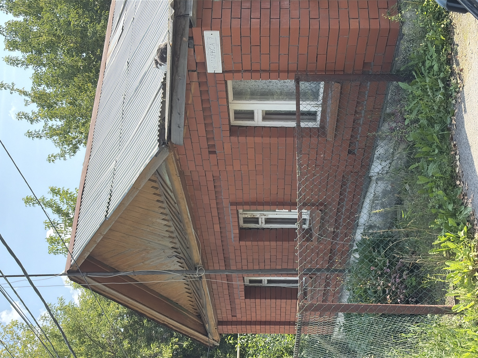 The image shows a brick house with a corrugated metal roof. A street sign reading "К. МАРКСА 7" is visible on the corner of the house. There are two windows on the side of the house facing the street. A chain-link fence runs along the front of the property, with overgrown vegetation behind it. A gray and white cat is sitting on the roof of the house. Trees are visible in the background, and power lines run overhead. The scene appears to be in a residential area.