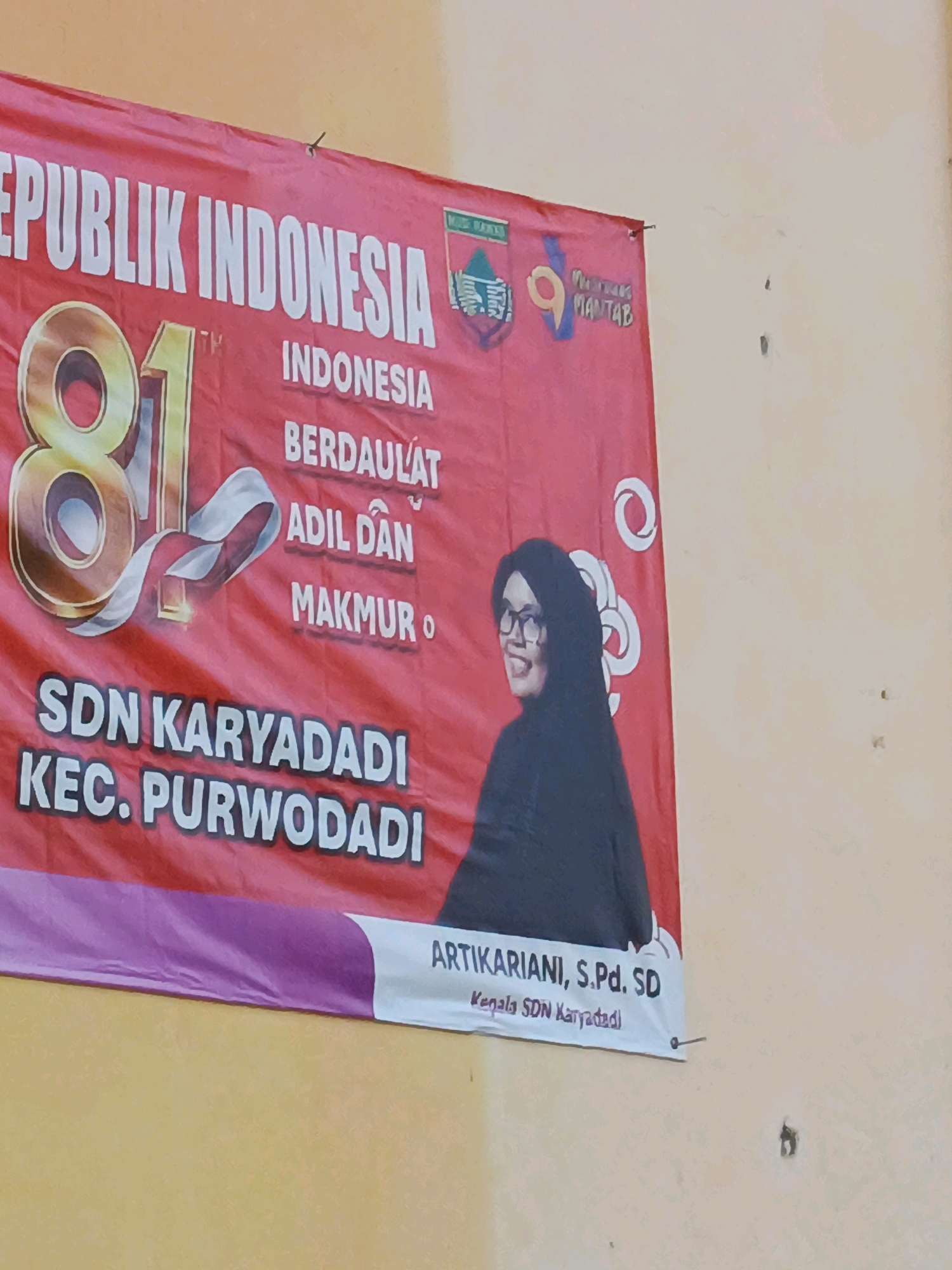 A banner hangs on a light-colored wall. The banner is red and displays text and graphics. At the top left of the banner, in large white letters, it reads "REPUBLIK INDONESIA". To the right of this text, there is a graphic of an eagle and the word "MANTAB" in smaller letters. Below "REPUBLIK INDONESIA" is a large number "81" in a golden gradient with a white ribbon entwined around it. Below the number, in white text, it says "INDONESIA BERDAULAT ADIL DAN MAKMUR O". On the bottom left of the banner, in large white letters, it reads "SDN KARYADADI KEC. PURWODADI". To the right of this text, a woman wearing a black hijab and glasses is depicted. The bottom right of the banner has a white section with text that reads "ARTIKARIANI, S.Pd. SD Kepala SDN Karyadadi". The banner is attached to the wall with small nails. The wall appears to be textured and has a few small dark marks on it. The scene is indoors, likely a school or community building, given the text on the banner identifying it as "SDN KARYADADI KEC. PURWODADI".