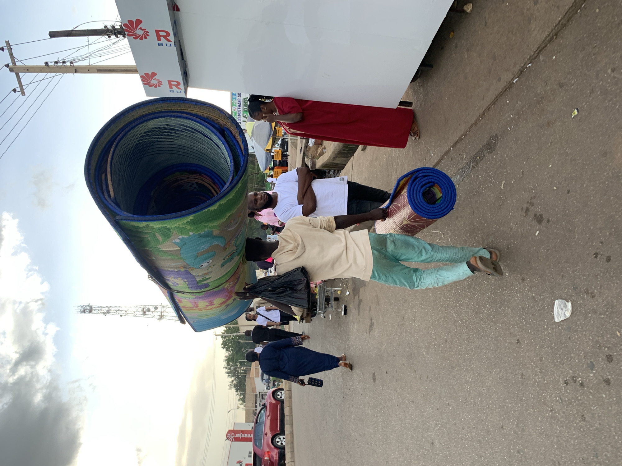 In Jos, Nigeria, a man in a light brown hoodie and teal pants carries a large rolled-up mat on his head, while another rolled mat is slung over his shoulder. He walks on an asphalt road, which appears to be a busy street with other pedestrians and vehicles. To the left, a woman in a blue dress with a patterned handbag walks away from the camera, and a red car is parked on the side of the road. To the right, a large white structure, possibly a stall or a truck, is visible, with a woman in a red dress standing beside it. The background features a telecommunications tower, power lines, and a cloudy sky, indicating an outdoor urban environment. The overall scene depicts everyday life and commerce in the city.