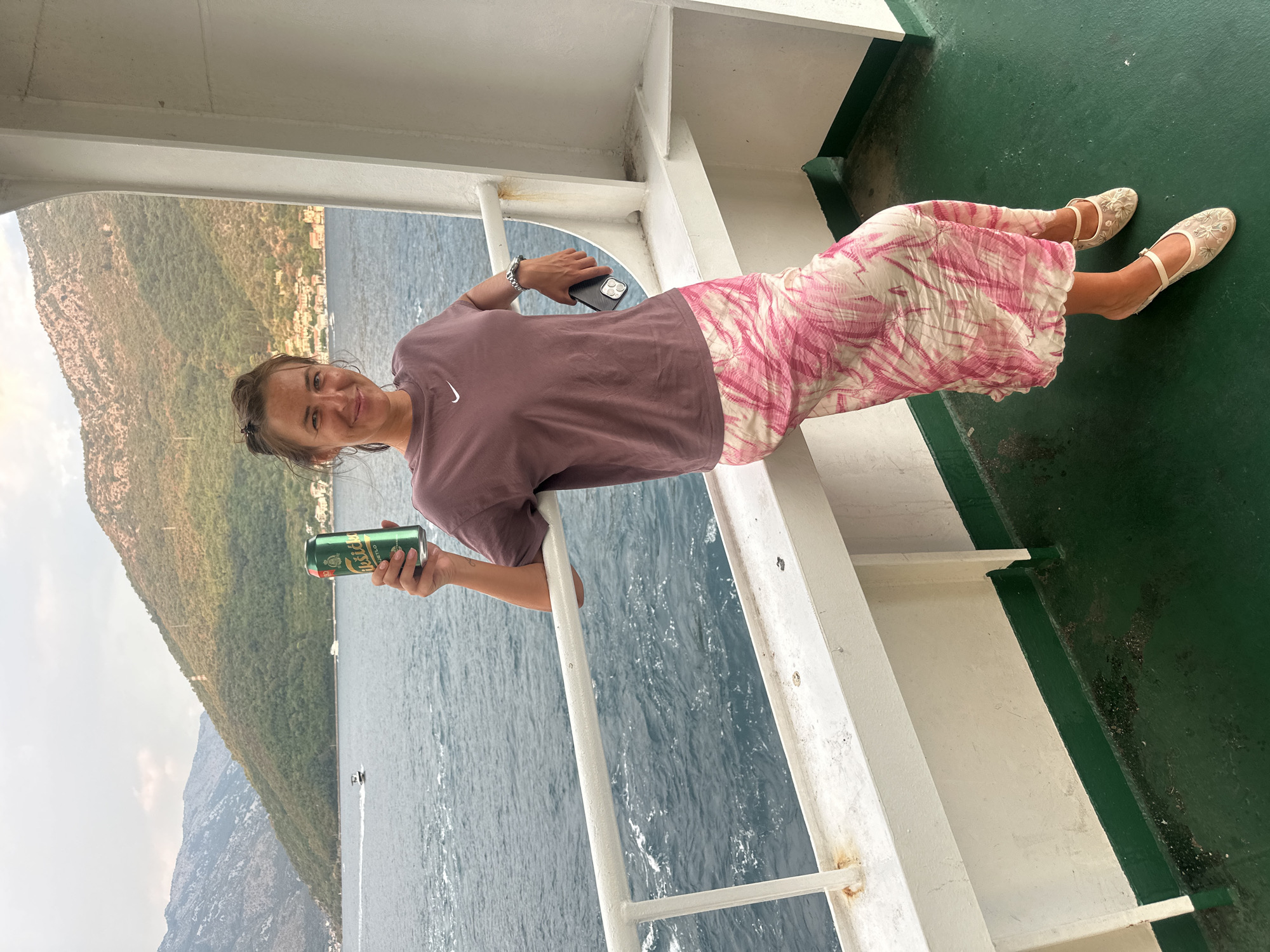 A woman stands on the deck of a ferry, leaning against a railing. She holds a green can of Nikšićko beer in her right hand and a black smartphone in her left. She is wearing a dusty rose t-shirt with a white Nike logo and a long, flowing skirt with a pink and white tie-dye pattern. Her feet are adorned with delicate, flat shoes with floral embroidery. Behind her, a calm, blue body of water stretches towards a coastline lined with buildings and verdant mountains. The sky is overcast. The ferry deck is painted green, with a white railing and a white bulkhead behind the woman. The scene is set in the Bay of Kotor, near the village of Đurići, Montenegro.