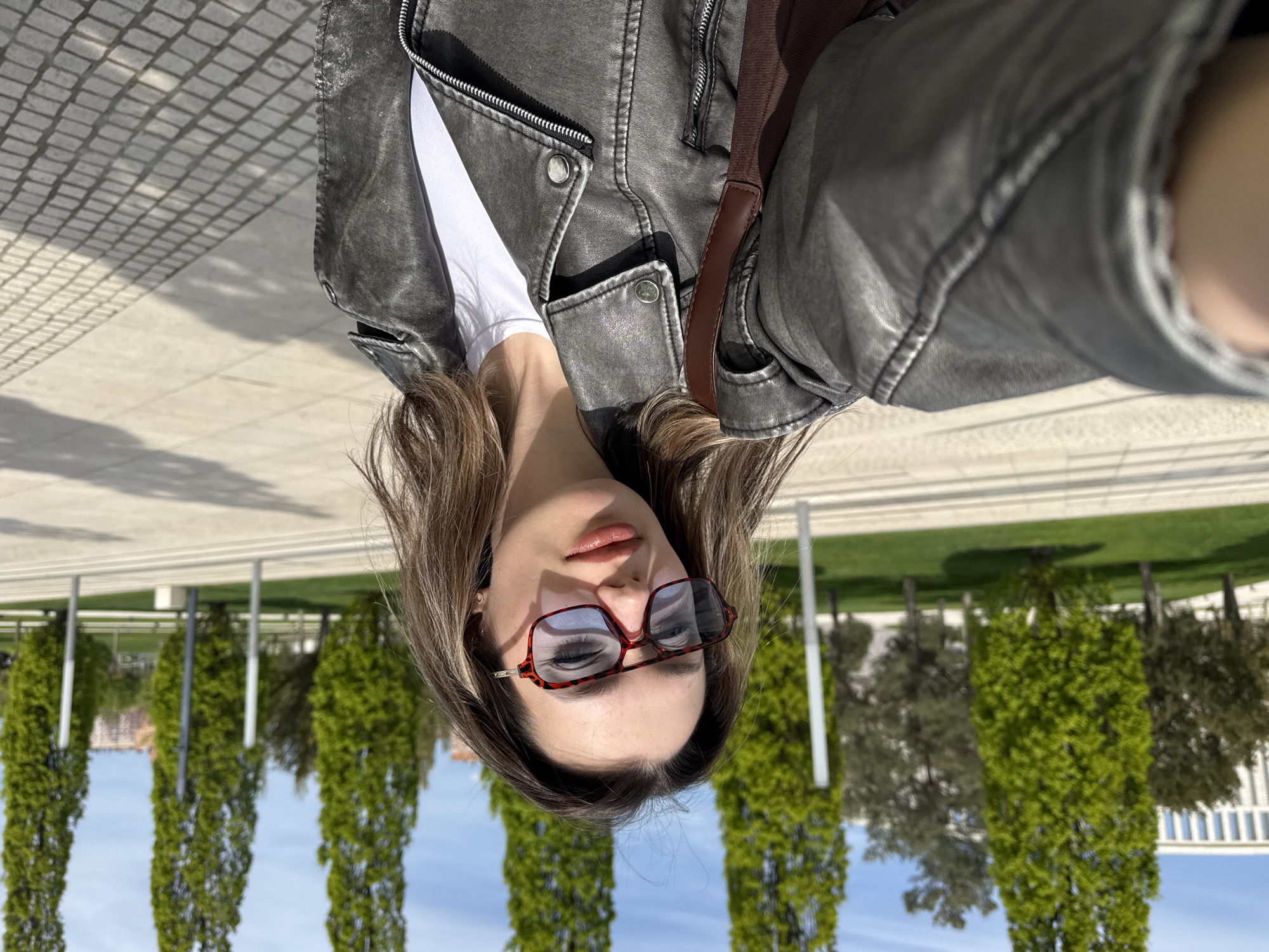 The image is a selfie of a young woman taken outdoors on a bright, sunny day. She has medium-length brown hair and is wearing a dark grey, distressed-look leather-style jacket over a white t-shirt. A brown strap, likely from a bag, is visible over her right shoulder. She wears oversized sunglasses with distinctive red or tortoiseshell frames and subtly tinted lenses, which partially obscure her eyes. Her lips are a soft coral shade, and her expression is neutral.
The background depicts a well-maintained outdoor public space, possibly a park or square. The ground features varied paving, including a textured, cobblestone-like area with distinct shadows indicating strong overhead sunlight, and smoother, lighter-colored tiles. In the mid-ground, a neat row of tall, slender, green trees, resembling cypresses, stands against a clear blue sky. Further in the distance, more green foliage and some light-colored architectural structures or railings are visible. No visible text is present in the image.
