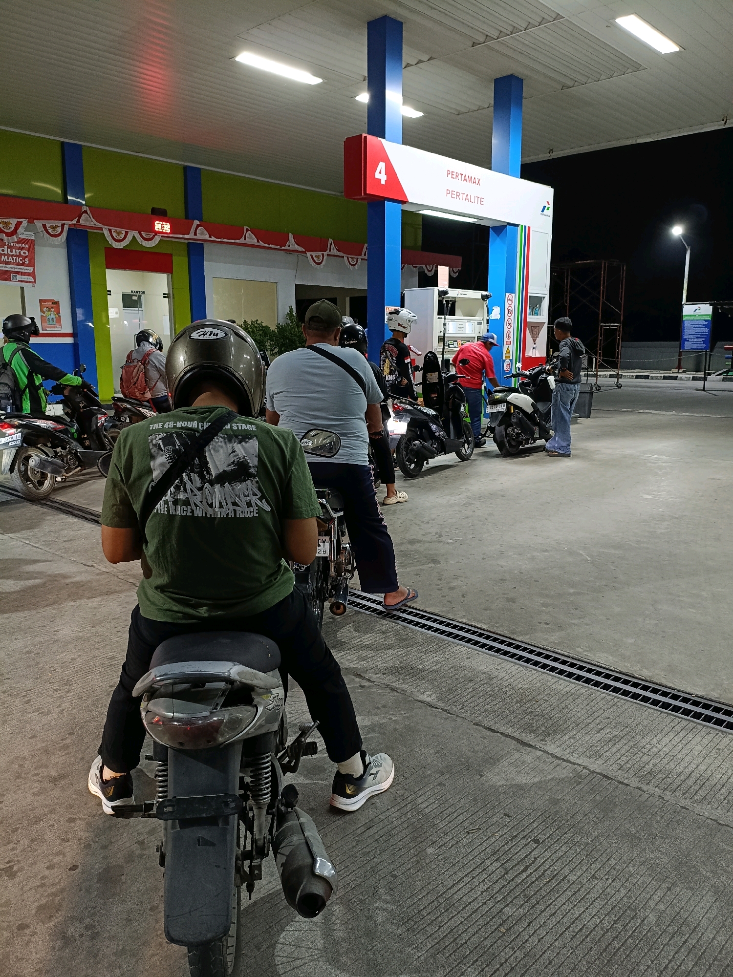 The image captures a nighttime scene at a Pertamina gas station in Maguwoharjo, Indonesia. Several motorcycles and their riders are present, either waiting or fueling. In the foreground, a person wearing a green t-shirt and black pants is seated on a motorcycle, facing away from the camera. The t-shirt has a graphic print. Behind this individual, other people are visible, some also on motorcycles, and one person in a red shirt appears to be attending to a motorcycle. The gas station features bright lighting, with a prominent sign indicating "PERTAMAX PERTALITE" and a large number "4." The ground is paved concrete, with visible drainage grates. The overall atmosphere suggests a typical evening at a busy fuel station.