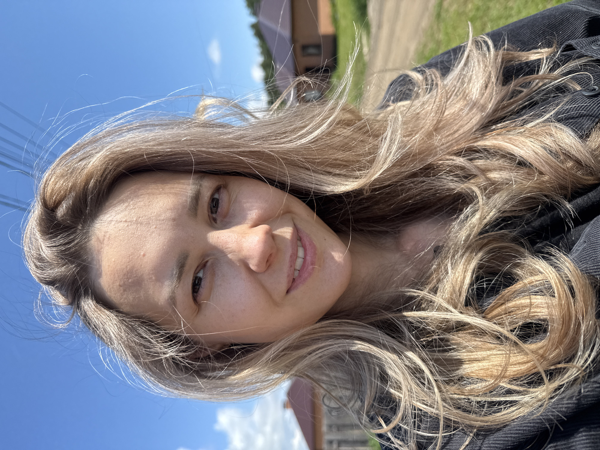 This is a bright, sunlit outdoor selfie of a woman in Chaikovsky, Russia. The woman, appearing to be young to middle-aged, is positioned in a close-up, chest-up shot, smiling gently at the camera with her head slightly tilted. She has long, wavy, blonde and light brown hair with visible highlights, which appears wind-blown and catches the strong natural sunlight, creating a dynamic effect. Her smile is warm and natural, revealing some teeth, and her eyes are light brown. The strong sunlight illuminates her face, highlighting her features and creating some shadows. She is wearing a dark-colored, possibly denim or corduroy, jacket or shirt with visible buttons on the front.
The background features a vibrant blue sky with a few wispy white clouds, indicative of a clear and sunny day. Power lines are visible against the sky on the left side of the frame. In the blurred mid-ground, there are suggestions of distant buildings, possibly houses with reddish roofs, and green foliage, indicating a rural or suburban outdoor setting. A dirt path or road is also faintly visible in the background. The overall impression is cheerful and natural, capturing a moment of contentment under bright sunshine. There is no visible text in the image.