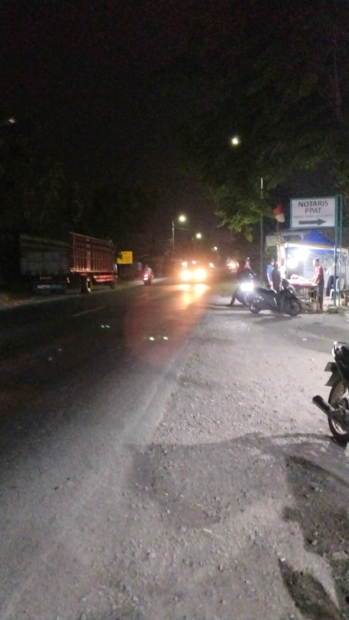 This image captures a nighttime street scene in Canggu, Indonesia. A red and silver truck is parked on the left side of the road, facing right. Further down the road, the headlights of oncoming vehicles are visible, creating a bright glare. On the right side of the street, a small outdoor stall is set up with a blue tarp roof. A sign above the stall reads "NOTARIS PPAT" with an arrow pointing to the right. Three people are gathered near the stall, with two standing next to a motorcycle and another person attending to something at the stall. A motorcycle is parked on the far right of the frame, its rear wheel and exhaust pipe visible. The road surface appears to be paved with gravel and asphalt, and the scene is illuminated by streetlights and vehicle headlights. The overall atmosphere suggests a lively, possibly commercial area, even at night.