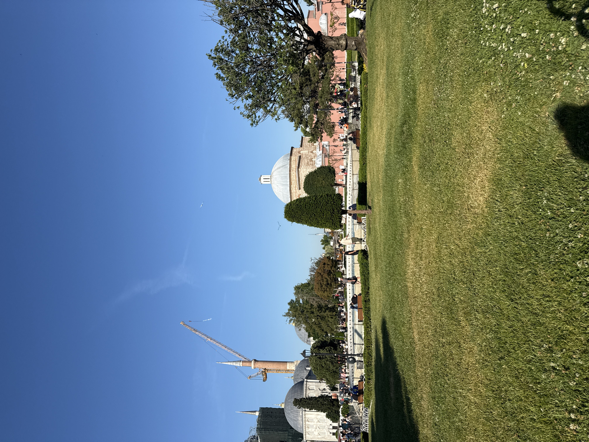 This image captures an outdoor scene at Sultanahmet Park in Istanbul, Turkey, on a clear, sunny day.
The background on the left features a large mosque, likely the Blue Mosque, with its upper sections and a minaret partially obscured by green scaffolding and netting, indicating renovation work. A prominent, tall, light brown construction crane stands adjacent to this minaret, its boom extending upwards. To the right of the mosque, another historic domed building with lighter colored walls is visible. In the mid-right background, a large domed building with pale pink-orange walls stands, consistent with a traditional Ottoman structure.
The mid-ground is defined by a wide, rectangular water feature or fountain with white stone coping, stretching horizontally across the scene. Several dark green park benches are positioned around this feature, occupied by various individuals. Multiple dark streetlights with ornate lamps are also present along the fountain's edge.
Numerous people are dispersed throughout the scene. Some are walking on paved pathways behind the fountain, while others are seated on the benches. Their attire is casual. Small, manicured green hedges line the fountain structure.
The foreground is dominated by a large expanse of green grass, which shows varying shades and some drier, yellowish patches. Small white clover flowers are visible throughout the grass. Dark shadows, likely cast by the photographer and other unseen objects, appear in the bottom-left and bottom-right corners of the frame.
The sky above is a clear and vibrant blue, with a faint, wispy contrail visible in the upper-mid section. Several small dark specks, likely birds, are scattered across the sky, and one larger white bird is present in the mid-left sky.