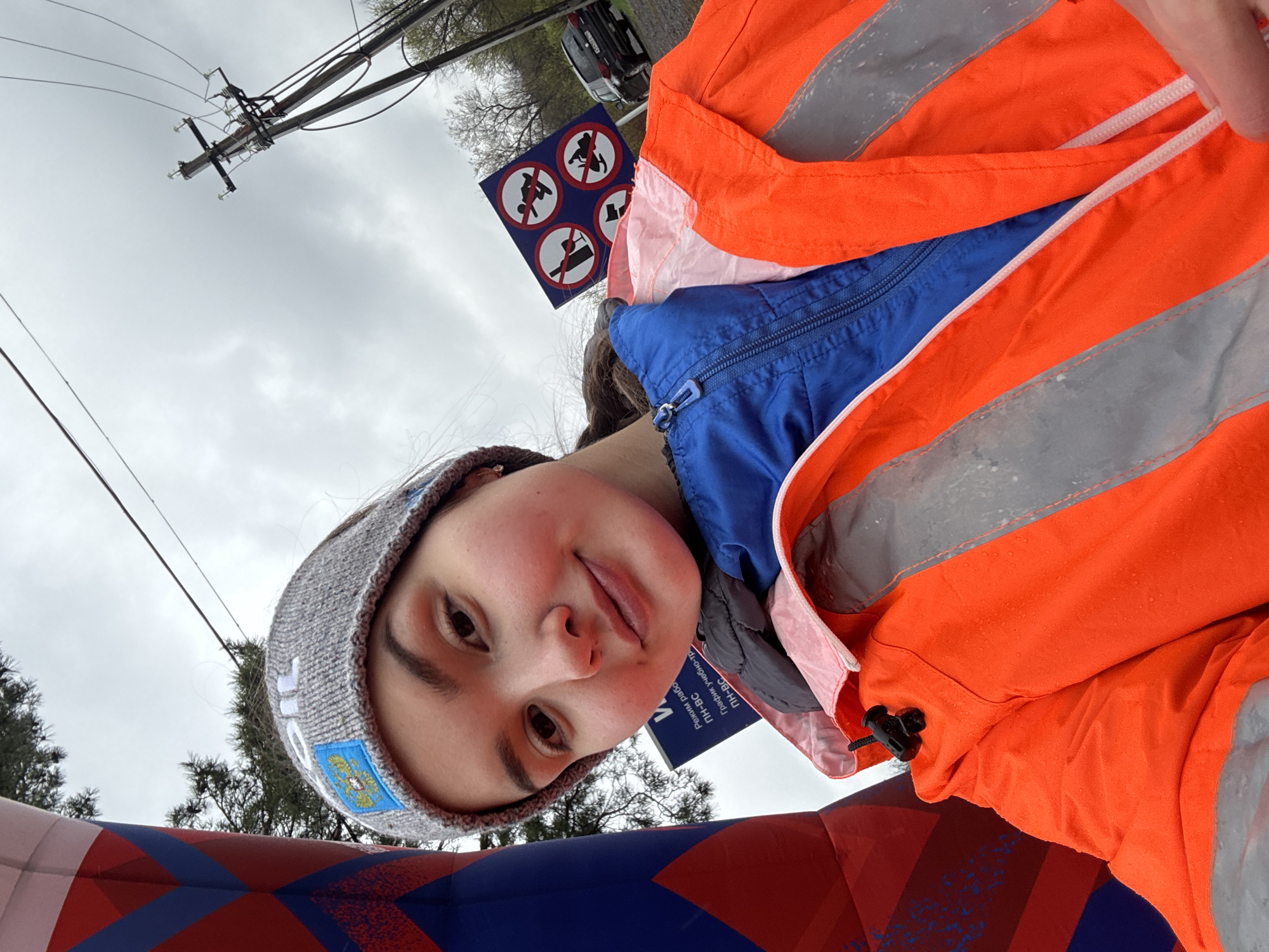 A person wearing a grey beanie with a blue emblem and an orange safety vest is posing for a selfie in front of a blue and red inflatable arch. Behind the person, there is a sign with several symbols indicating prohibited activities. An electrical pole and overcast sky are also visible. The location is presumed to be in Orenburg, Russia.
