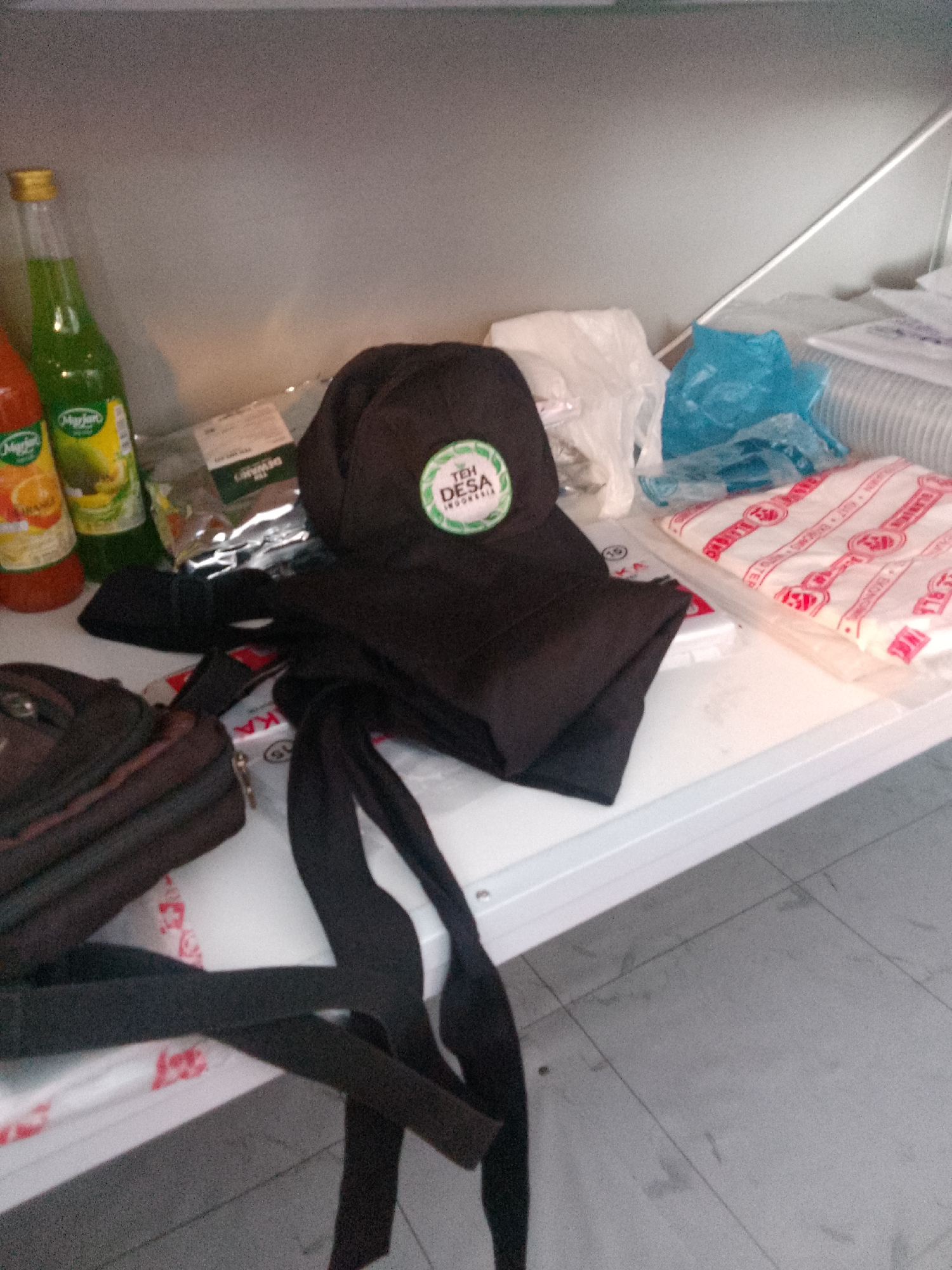 A shelf is cluttered with various items. On the left, two bottles of what appear to be flavored syrup are visible, one orange and one green. Next to them are several packets of tea, one of which is labeled "TEH DESA INDONESIA" with a circular logo. A black baseball cap with the same logo is placed on top of a folded black garment, likely a shirt or jacket. To the right of the tea packets, there are several plastic-wrapped items, including a blue plastic bag and some white packaging. In the foreground, a dark brown bag rests on a white surface. A black strap or tie is draped across the front of the shelf and extends to the left. The shelf itself is a light-colored, possibly white, material. The background appears to be a plain, light-colored wall. The floor below the shelf is tiled in a gray pattern. The overall scene suggests a storage area or a display shelf.