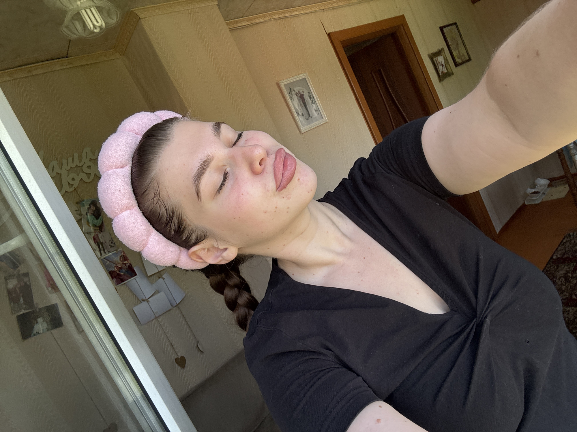 A selfie-style image shows a fair-skinned woman with a pink headband. She has dark hair in a braid. Her eyes are closed and she is pouting. She is wearing a black V-neck shirt. The background includes a window, wall with pictures, and a partially visible doorway. Location is not discernible from image.