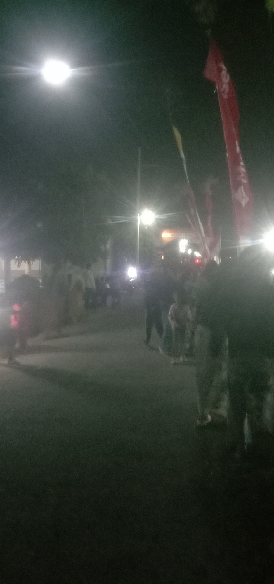 The image depicts an outdoor night scene in what appears to be a street in Banjar Dowo, Indonesia. Several individuals, including adults and children, are visible, some standing and others blurred due to movement or low light conditions. The environment is dimly lit by streetlights, one of which is prominently bright in the upper left portion of the frame. There are flags present, with a large red flag being noticeable on the right side, bearing some text that is difficult to discern. The ground appears to be asphalt, suggesting a road or paved area. The overall atmosphere is somewhat hazy, possibly due to the night, lighting, or camera quality.