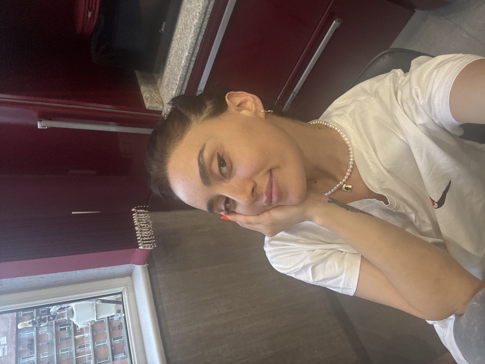 A young woman with dark hair pulled back and adorned with a pearl necklace is taking a selfie in what appears to be a kitchen or dining area. She is wearing a white t-shirt and resting her head on her hand. The background includes red cabinetry, a granite countertop, and a microwave. A window to her left reveals a view of a multi-story apartment building. The lighting suggests it is daytime. The overall impression is a casual, indoor portrait.