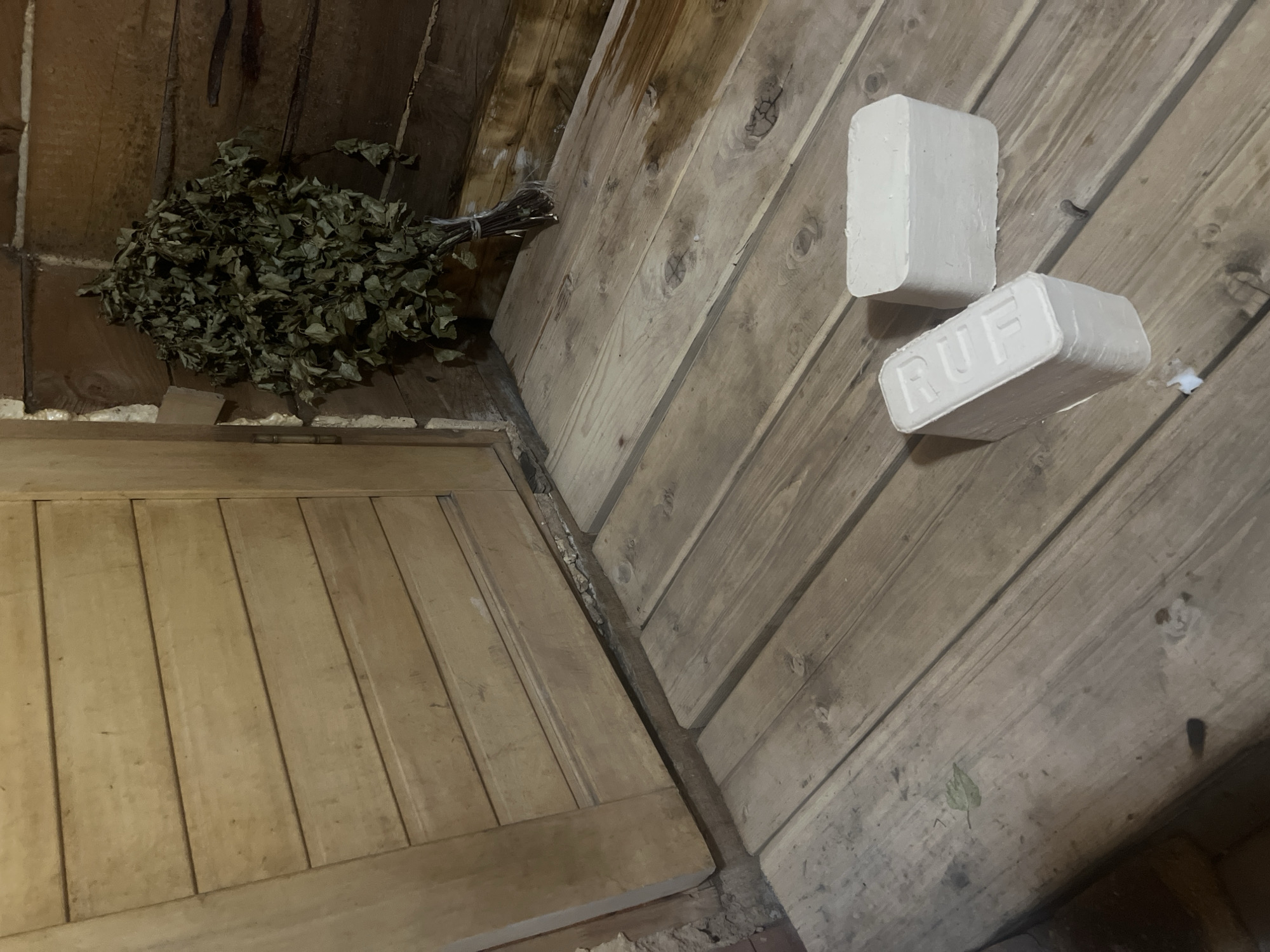 A medium-shot depicts the interior of a likely sauna, featuring wooden construction. In the upper-left corner rests a bundle of dried birch branches for traditional sauna use. Two white RUF wood briquettes are situated diagonally in the upper-right, against the wooden wall. Below the birch branches is a constructed-in wooden bench. The image was likely taken in a sauna in Chaikovsky, Russia.