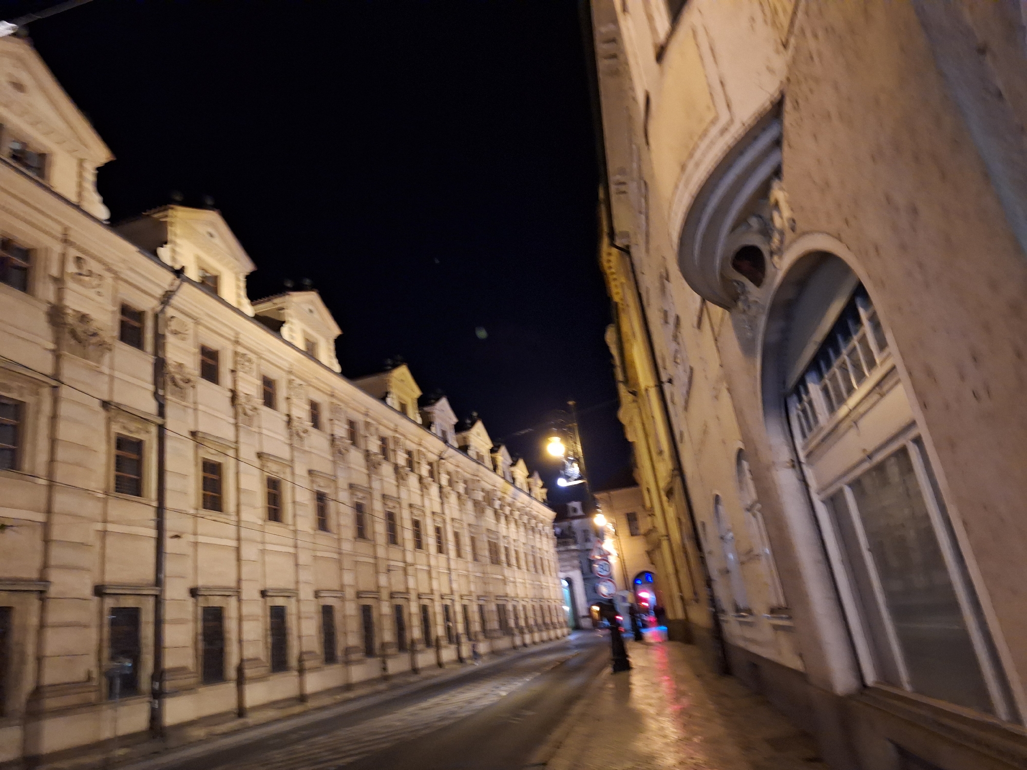 The image depicts a street scene at night in Prague, Czechia. A long, multi-story building with a light-colored facade and numerous windows lines the left side of a paved street. The building exhibits architectural details such as cornices, decorative elements above windows, and stepped gables. On the right side, another building with a light facade is partially visible, featuring a curved architectural element and large windows. Streetlights illuminate the scene, casting reflections on the wet street surface. A lone figure is seen walking on the right side of the street. Traffic signs, including a circular sign with the number "30", are visible in the distance. The overall impression is that of a historic European city at night, with the buildings and street lit by artificial light.