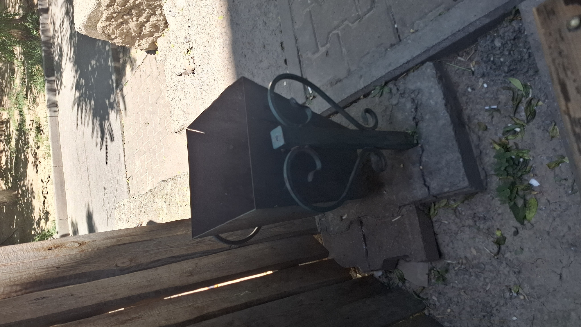 The image depicts an outdoor scene, likely in Shymkent, Kazakhstan, showing a dark metal trash receptacle with decorative scrollwork on its stand. The receptacle is positioned on a base of concrete slabs, which are placed on the ground composed of dirt and scattered green leaves and cigarette butts. To the left of the trash receptacle is a weathered wooden fence. In the background, a paved area, possibly a sidewalk or road, is visible, with trees casting shadows. The environment appears to be public, with evidence of human presence such as the discarded cigarette butts.
