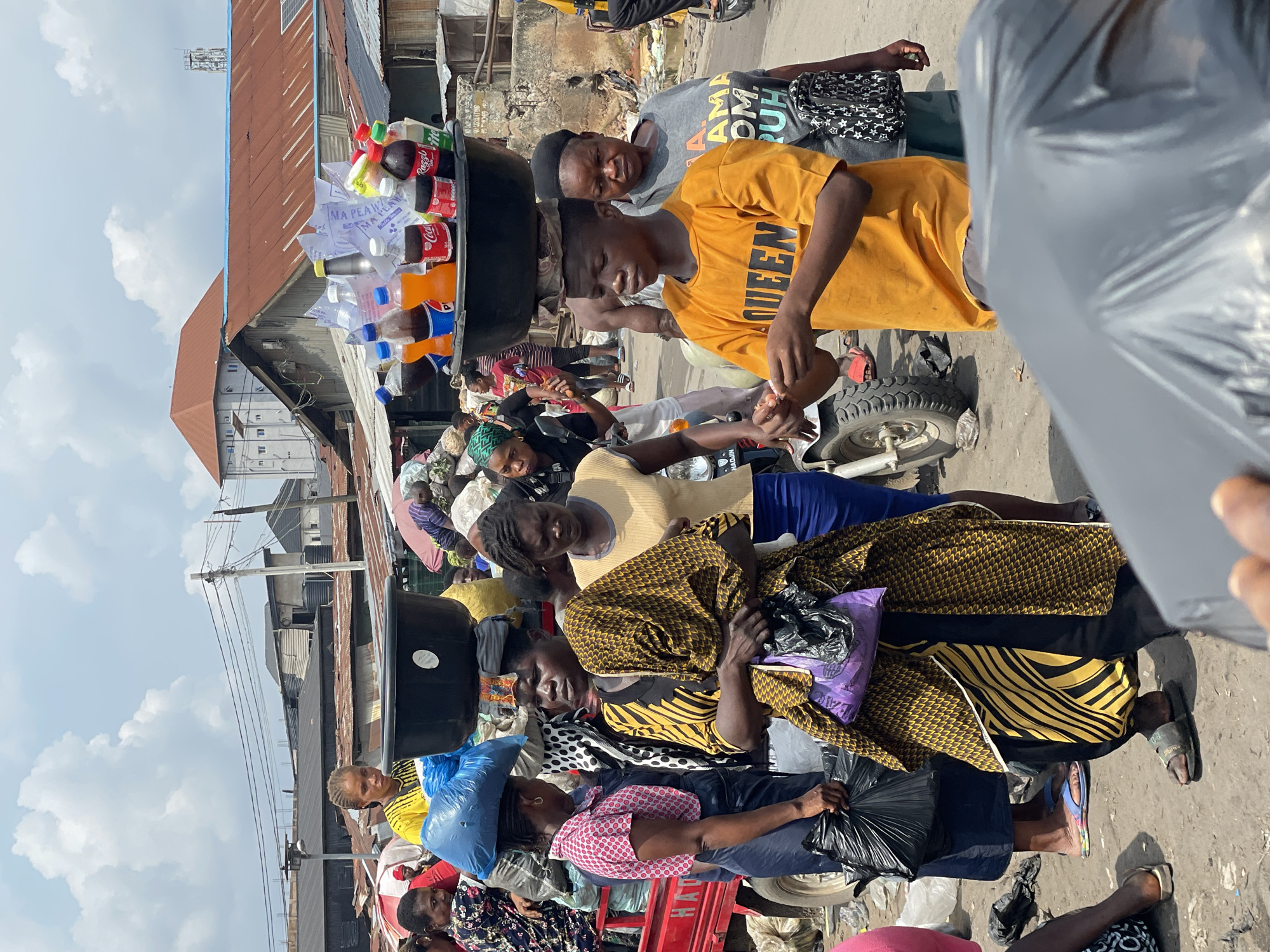 This media file captures a bustling outdoor scene, likely a market or busy street in an urban environment, under bright daylight with a partly cloudy sky. The image features numerous people, mostly adults, engaged in daily activities.
A central figure is a young person in a yellow t-shirt, carrying a large black basin on their head filled with various bottled drinks, including Coca-Cola, Fanta, and water bottles. Text on the yellow t-shirt reads "QUEEN," while another person in a grey t-shirt has partially visible text that appears to be "AMAZON." A white plastic bag among the drinks has "MA PEAWA" printed on it. Many individuals are seen carrying plastic bags of various colors (blue, black, purple), some on their heads.
In the background, weathered buildings with corrugated metal roofs and utility poles with wires are visible. Some roofs have small solar panels. A partial view of a small vehicle, possibly a motortaxi, is also seen. The people's attire and the general infrastructure suggest a location in a developing country. The scene portrays typical street commerce and daily life, with people walking and carrying goods. Faces are generally neutral or focused on their activities.