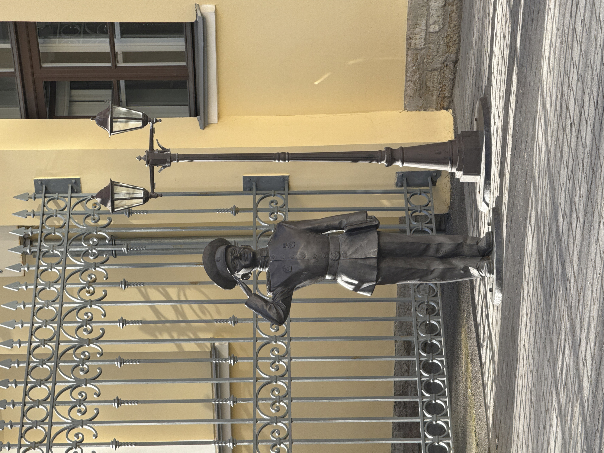 An outdoor, rotated vertical photograph captures a bronze statue of a man in a military-style uniform saluting, positioned in front of a decorative metal gate in Saint Petersburg, Russia.
The statue depicts a man wearing a peaked cap, a double-breasted jacket adorned with buttons and possibly medals, and trousers. His right hand is raised to his cap in a formal salute, while his left arm is bent at the elbow, hand resting near his waist. He has a mustache and a focused expression. The statue stands on a dark, possibly stone, base.
Behind the statue is an ornate gray metal gate or fence featuring spear-like tops, circular and scroll patterns, and twisted vertical bars. Beyond the gate, a light yellow building wall is visible, with a dark-framed window towards the upper left. To the right of the statue stands a classic-style dark street lamp with two lanterns, its base partially obscured by the statue.
The ground is paved with what appears to be patterned cobblestones or concrete slabs, creating a grid-like texture. Long, distinct shadows are cast across the pavement and through the gate, indicating a bright, sunny day, likely during the morning or late afternoon. No text is visible in the image. The scene suggests a public space, possibly a memorial or a historical site.