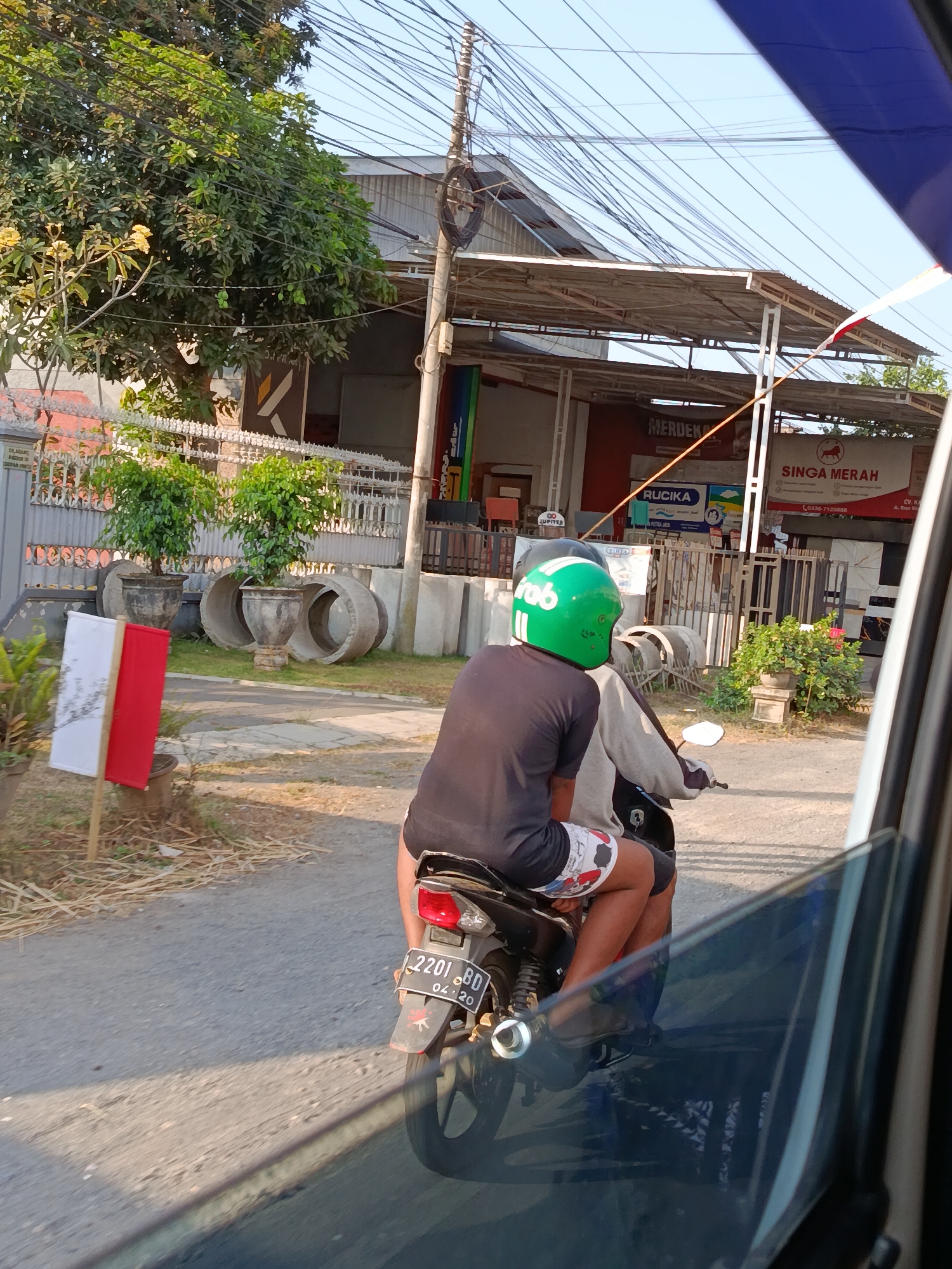 A person wearing a green helmet with a white logo is riding a motorcycle with another person as a passenger. The driver is wearing a dark shirt and patterned shorts, while the passenger is wearing a gray jacket. The motorcycle's license plate reads "2201 BD 04.20". They are on a paved road in front of a building with a corrugated metal roof. The building has several signs, including one that says "MERDEKA" and another with a lion logo and "SINGA MERAH". There are trees and plants around the building, as well as concrete pipes. The image appears to be taken from inside another vehicle, with part of a tinted window visible in the foreground. The setting is consistent with a street scene in Wonosari, Indonesia.