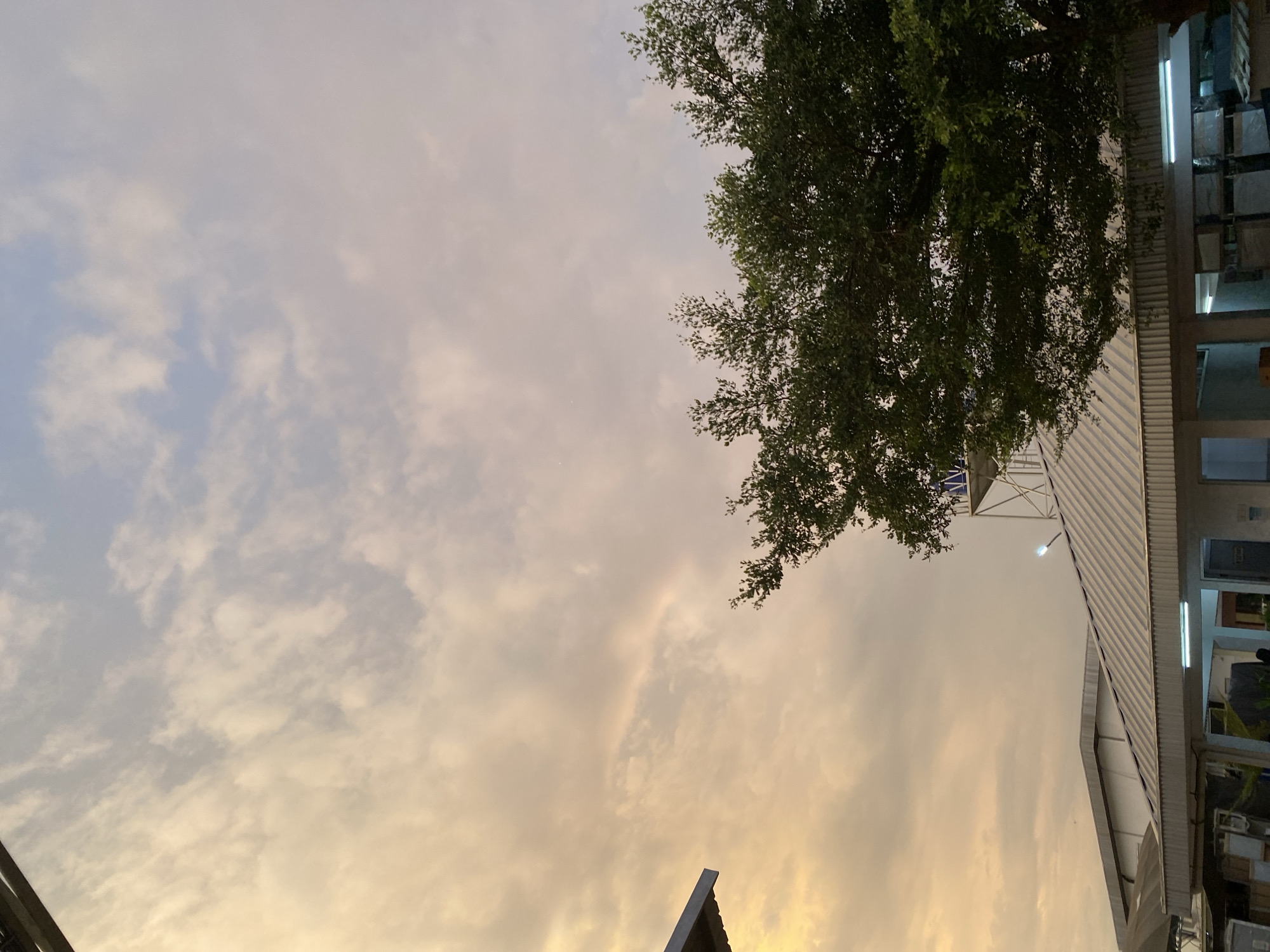 This is a photograph taken outdoors, likely in Gambut Barat, Indonesia. The image is oriented vertically, with the sky filling most of the frame. The sky displays a mix of light blue and warm, golden-orange clouds, suggesting it might be either sunrise or sunset. Wispy clouds are scattered across the sky, creating a soft and diffused light.
In the upper right quadrant of the image, a large tree with lush green foliage is visible, its branches reaching into the sky. To the right of the tree, a building with a corrugated metal roof is partially in view. The building appears to be a commercial or industrial structure, with several windows and what looks like shelving or display units visible inside. A bright fluorescent light is on inside the building, contributing to the ambient lighting.
A portion of a dark, angled structure, possibly a roof or awning, is visible in the lower left corner of the frame. No people are clearly discernible in the image. The overall impression is one of a tranquil moment captured at the transition between day and night, with a blend of natural and man-made elements. The warm tones of the sky evoke a sense of calm.