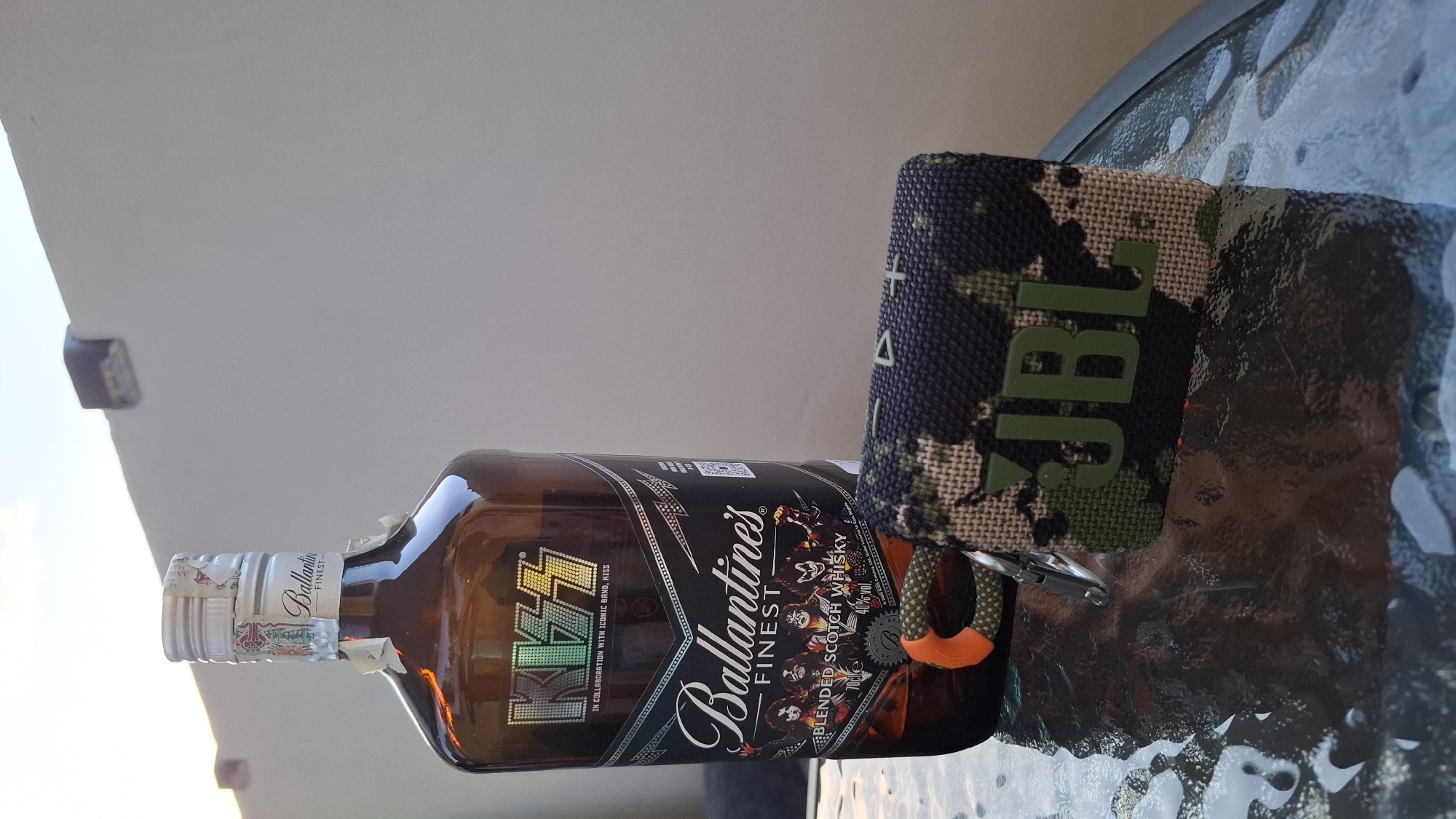 The image depicts a bottle of Ballantine's Finest blended Scotch whisky in a collaboration with the band KISS, lying on a glass-topped table outdoors. Next to the bottle is a JBL portable speaker with a camouflage pattern. The table appears to be on a balcony or patio, judging by the wall and ambient light. Without further information, it is impossible to verify that it is located in the city of Sofia, Bulgaria.