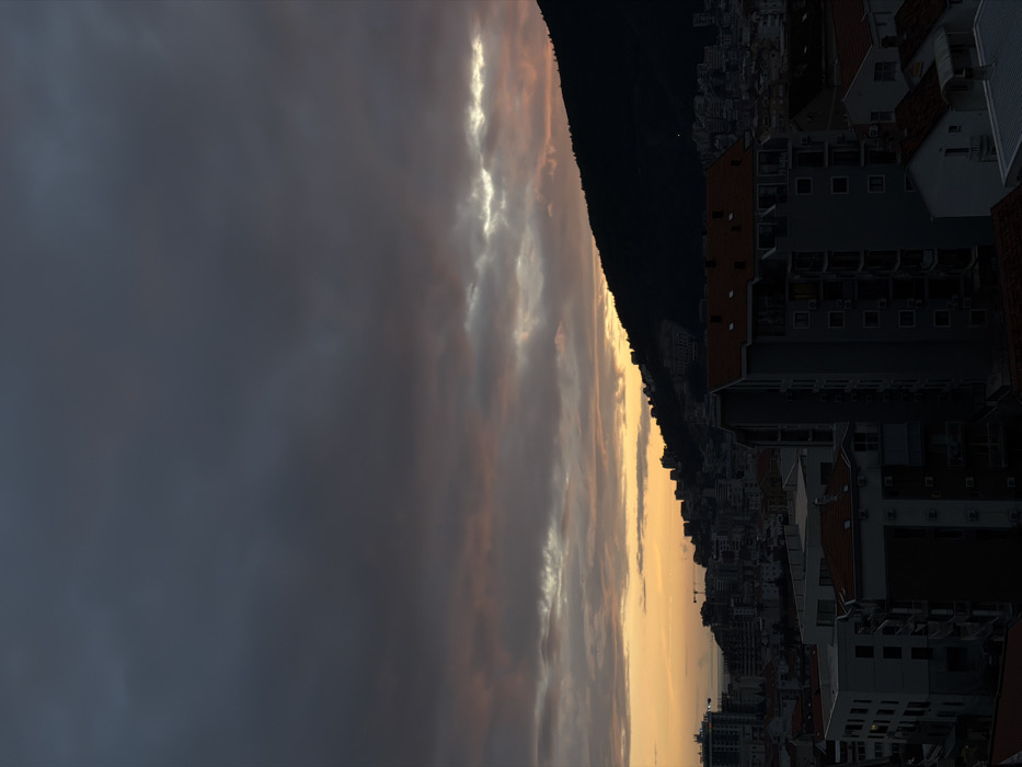 A panoramic view captures a dramatic sunset or sunrise over a coastal city, likely Budva, Montenegro, as suggested by the context. The sky dominates the upper portion of the frame, filled with a dynamic interplay of clouds. Dark, heavy grey clouds occupy the far left, while a central band features lighter, wispy clouds illuminated with warm golden and soft pink hues by the hidden sun. Bright, glowing streaks of light cut through the clouds, indicating intense sunlight.
Below the sky, a large, dark mountain or hill forms a prominent silhouette, its peak sloping down from left to right. Along the base of this mountain, and extending into the mid-ground, a dense urban landscape unfolds. Numerous buildings, including multi-story apartments and houses with distinctive red-tiled roofs, are visible. Most of these structures are cast in deep shadow, emphasizing the low angle of the sun. In the far right, a narrow strip of golden light beyond the buildings suggests the presence of the sea, reflecting the vibrant sky. The scene is devoid of any visible people or explicit activity, focusing instead on the atmospheric light and the urban-natural interface at twilight. No text is visible in the image.
