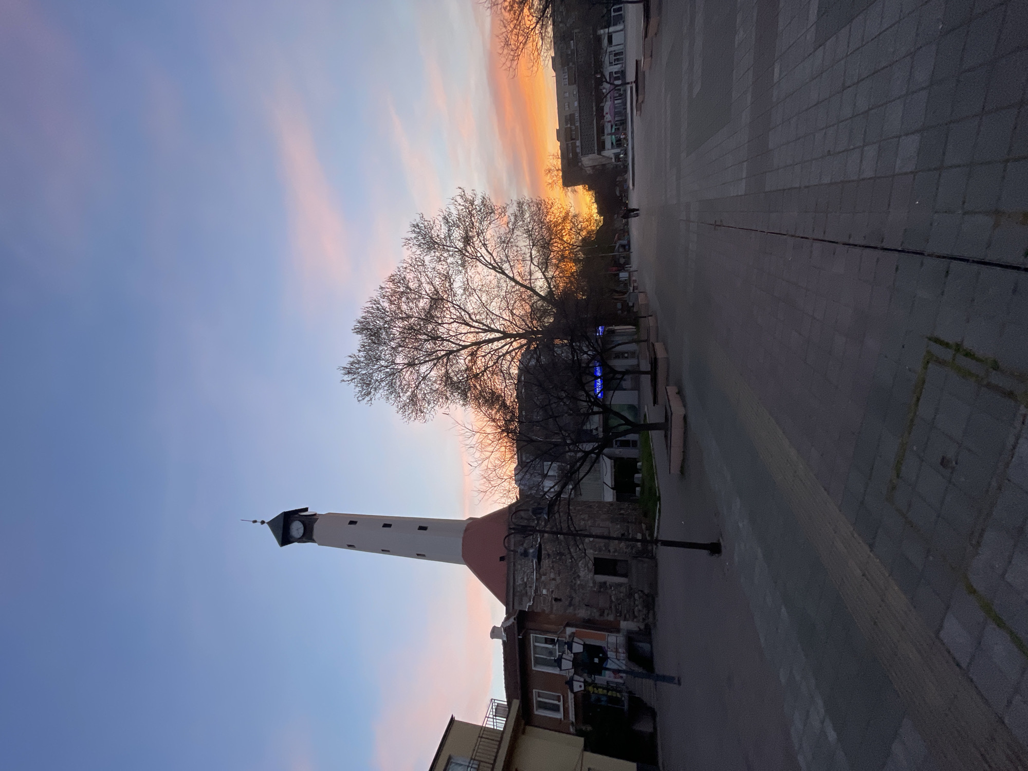 The image shows the Pazardzhik Clock Tower, located in the city of Pazardzhik, Bulgaria. The tower is constructed of stone and topped with a white clock tower with a black roof. The sky behind the tower is a mix of orange, pink, and blue, indicating either sunset or sunrise. A bare tree is situated near the tower, obscuring part of the building. The foreground features a paved square, and buildings can be seen in the periphery.