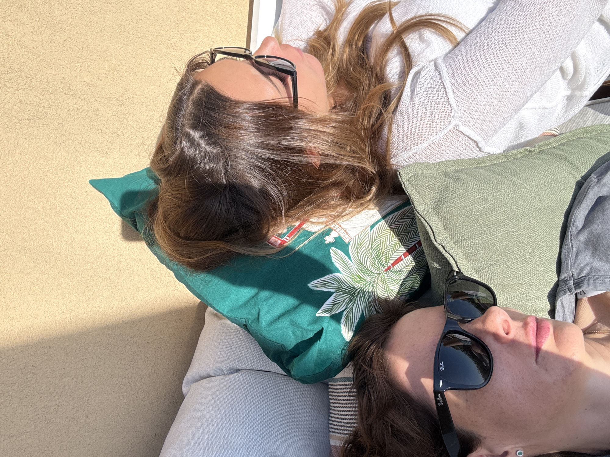 Two women are lying on cushions in an outdoor setting. One woman is wearing glasses and a white sweater, while the other has sunglasses on. The location cannot be determined based solely on the image.
