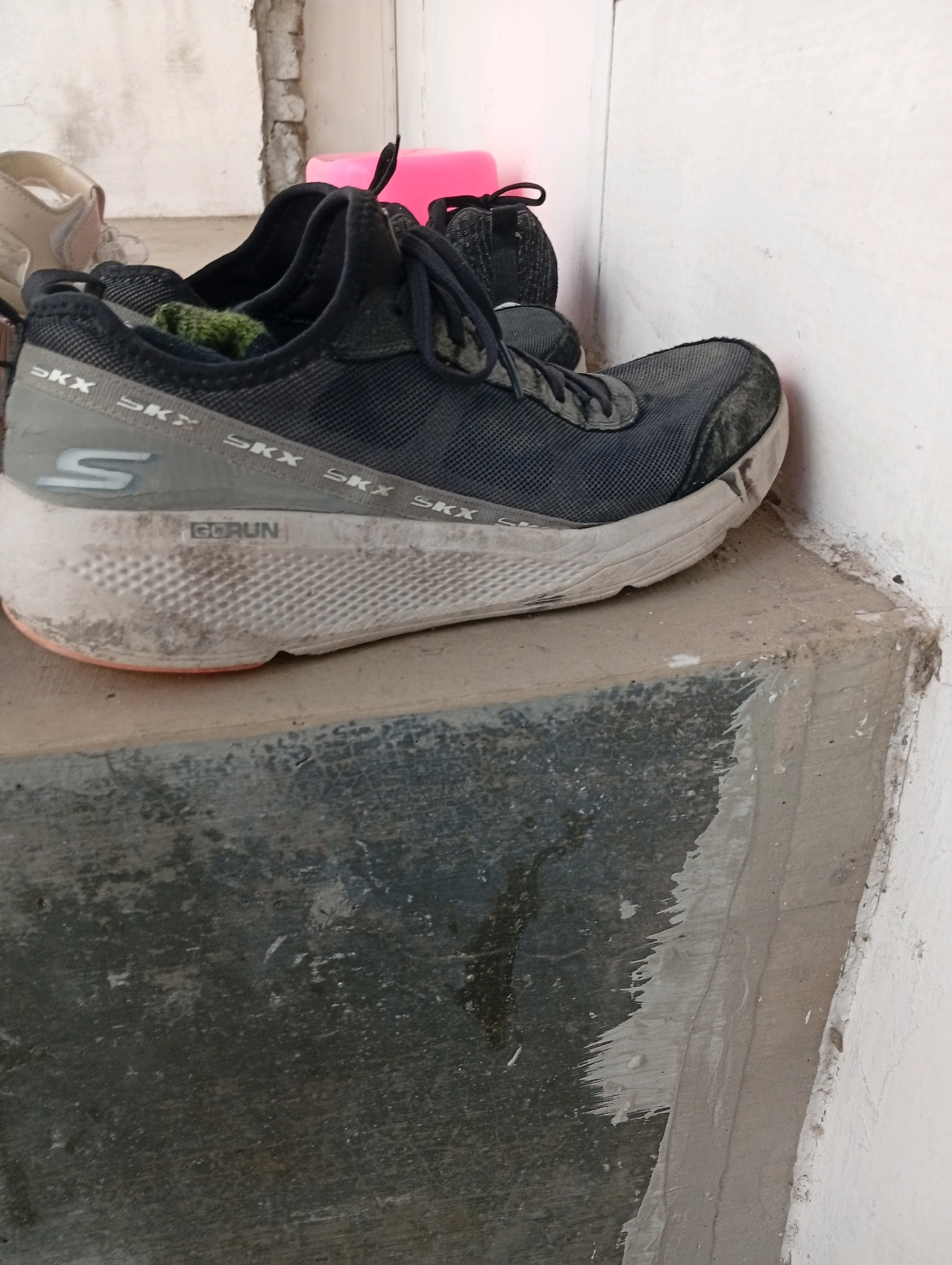 A pair of dark gray Skechers running shoes, with a white and orange sole, are placed on a concrete ledge in an outdoor setting. The shoes appear worn and dusty, with a green sock visible inside one of them. Behind the shoes, a portion of a white wall is visible, along with a pink object that might be a plastic container. The concrete ledge is stained and appears weathered. The scene is likely located in an urban or semi-urban environment, potentially a residential area in Banyu Ajuh, Indonesia, given the context provided, though no specific indicators of the city are visible in the image itself. The lighting suggests it is daytime.