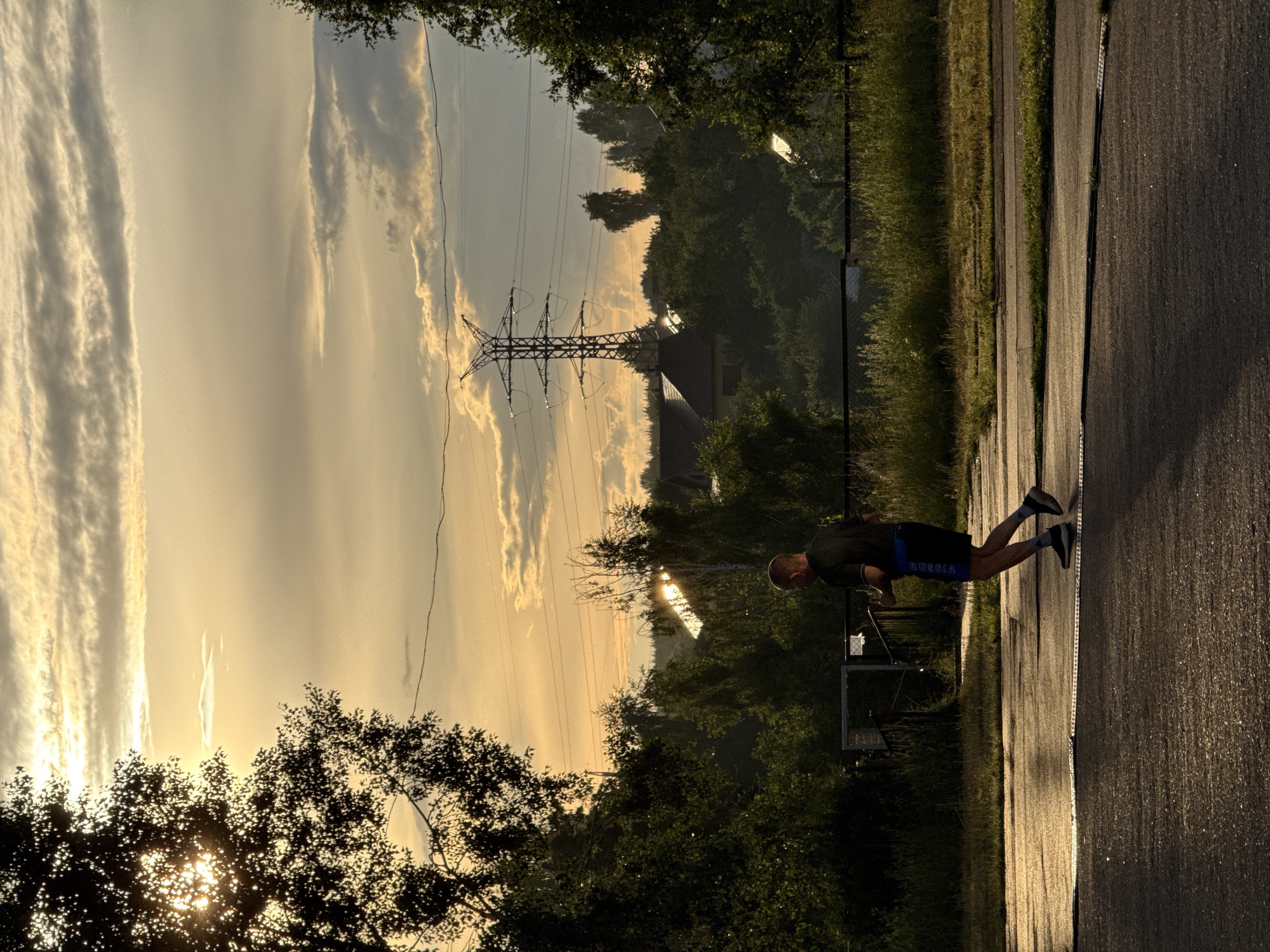 This is a vertical, low-angle, outdoor photograph taken at sunset in Chaikovsky, Russia. The setting is a paved path next to a grassy area with trees and a power line tower. The sky is filled with dramatic, wispy clouds, illuminated by the golden light of the setting sun, creating a warm and atmospheric glow. In the foreground, a man is performing push-ups on the paved path. He is wearing a dark t-shirt and blue shorts with white text that reads "RUSSIA". His body is in motion, suggesting he is engaged in an exercise routine. The overall mood is serene and active, with the natural beauty of the sunset contrasting with the human physical exertion.