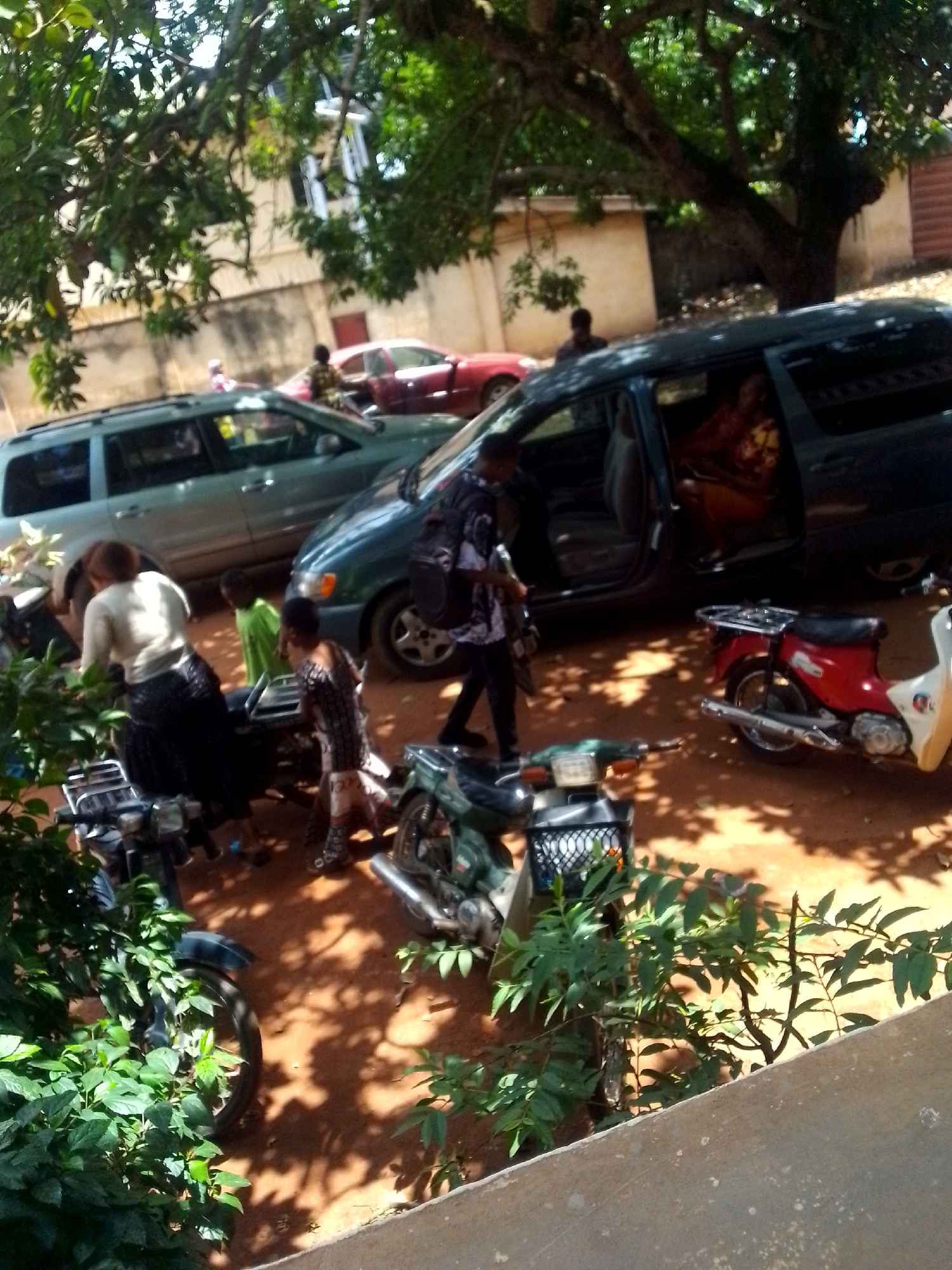 The image shows a gathering outdoors, seemingly in a residential area of Unknown, Unknown. Several people are present, including adults and children, interacting near a parked dark blue minivan. The vehicle's side door is open, revealing a seated individual inside. Another person with a shoulder bag is seen walking away from the van. Several parked motorcycles are scattered around the scene. The environment includes another vehicle, possibly a light blue SUV, parked nearby, as well as a red car further in the background. A building is visible behind the vehicles. The ground appears to be dirt or reddish soil, with foliage in the foreground and shadows cast by a large tree providing shade.