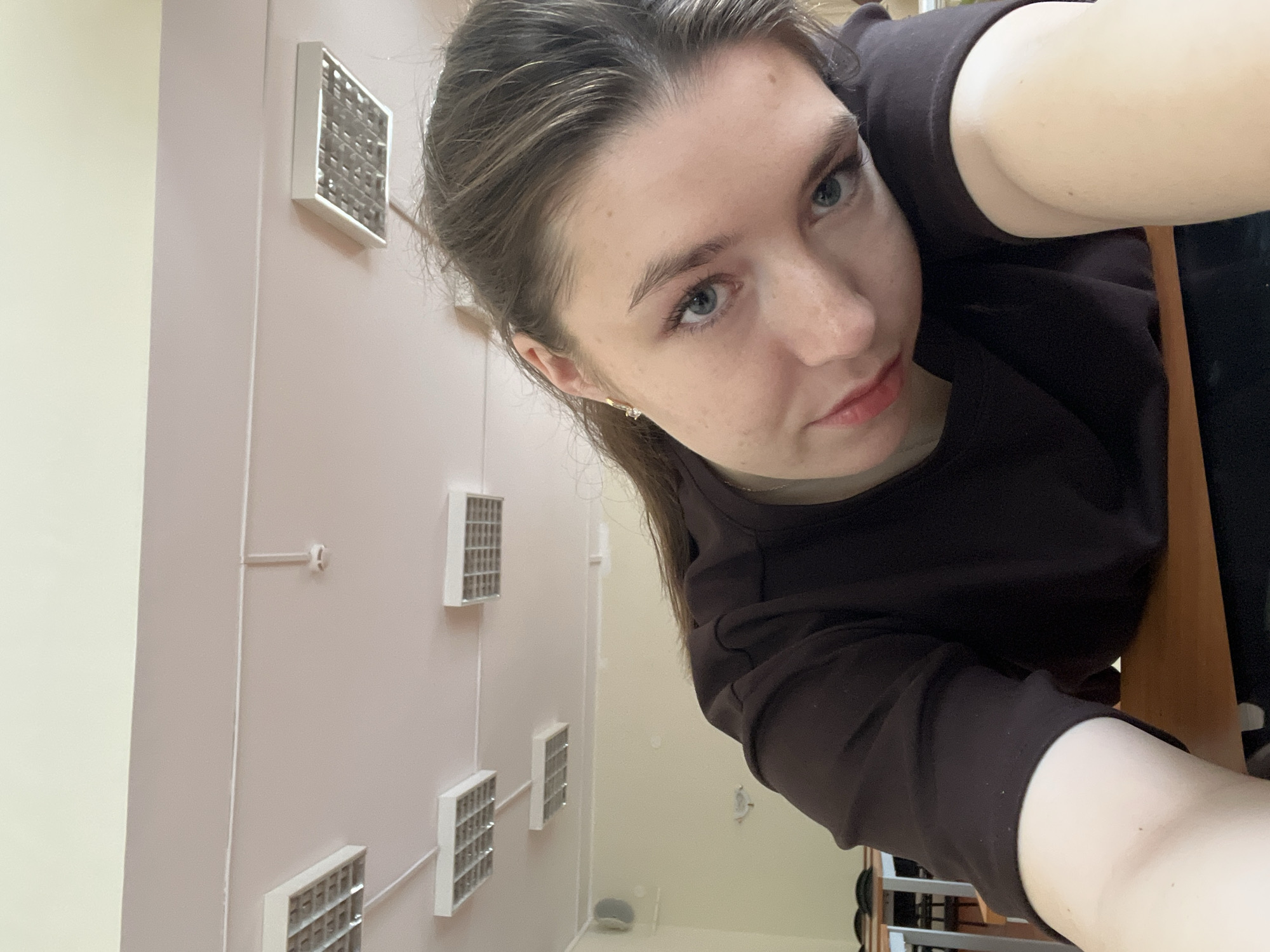 A woman with fair skin, blue eyes, and brown hair pulled back in a ponytail is shown in a selfie. She is wearing a dark brown top. The background features a light pink wall with a vertical row of square, white-framed air vents. A white wall is visible on the left. The location is implied to be a building in Khimki, Russia.