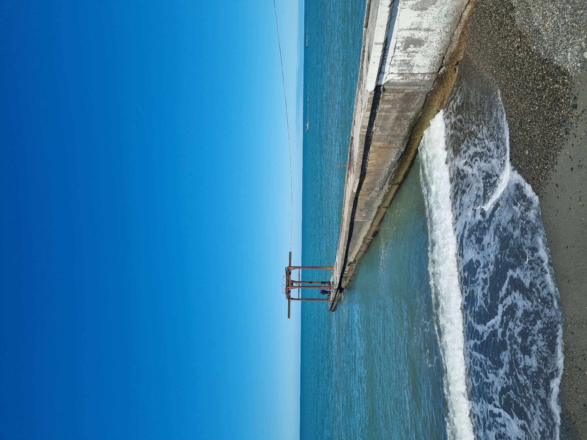 This media file captures a coastal scene, presented with an unusual vertical orientation where the horizon line runs vertically through the middle of the frame.
The left half of the image is dominated by a vast, clear, and vibrant blue sky, indicating bright, sunny weather. There are no clouds visible, and the sky's color graduates slightly from a deeper blue at the top left to a lighter tone towards the vertical horizon line. A thin, dark line, possibly a wire or cable, stretches horizontally across the sky and extends into the sea.
The right half of the image features the sea, a concrete breakwater or pier, and a narrow pebbly beach. The sea, a calm blue-green, occupies the space to the left of the breakwater. Gentle waves are breaking and receding along the shore, creating white foam that contrasts with the darker pebbles of the beach. The breakwater itself is a weathered, light-colored concrete structure, showing cracks and areas where the surface has eroded.
In the lower-middle portion of the image, extending from the breakwater into the sea, stands a rusty, brown metal structure, resembling an old frame, platform, or scaffolding. In the distant sea, along the vertical horizon, a few very faint and small shapes, possibly distant boats or vessels, are barely discernible.
The overall scene depicts a peaceful, sunlit coastal area, possibly a more utilitarian or older section of a beach or port, consistent with locations like Sochi, Russia. There are no people visible, and no discernible text appears in the image. The time of day appears to be mid-day or early afternoon due to the bright, direct sunlight.
